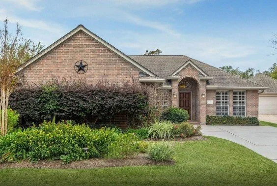 Brick house with a dark wooden front door, covered porch, and a star decoration on the upper front wall. Landscaped yard with bushes, grass, and plants.