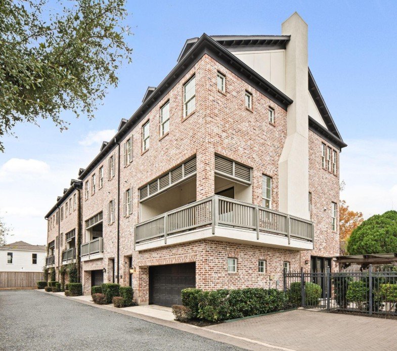 Multi-story brick apartment building with balconies, a black garage door, and gated entrance, surrounded by small bushes and trees.