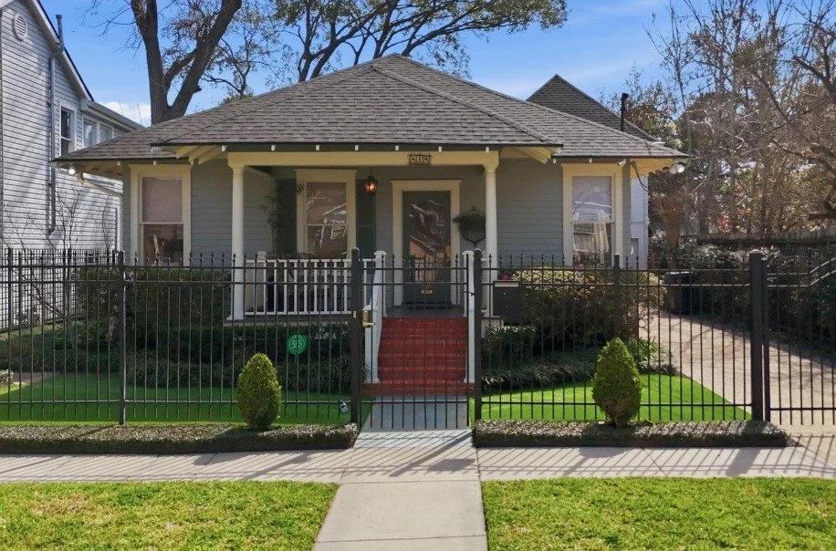 Front view of a small, light blue house with a porch, red carpet steps, fenced yard, and surrounding greenery.