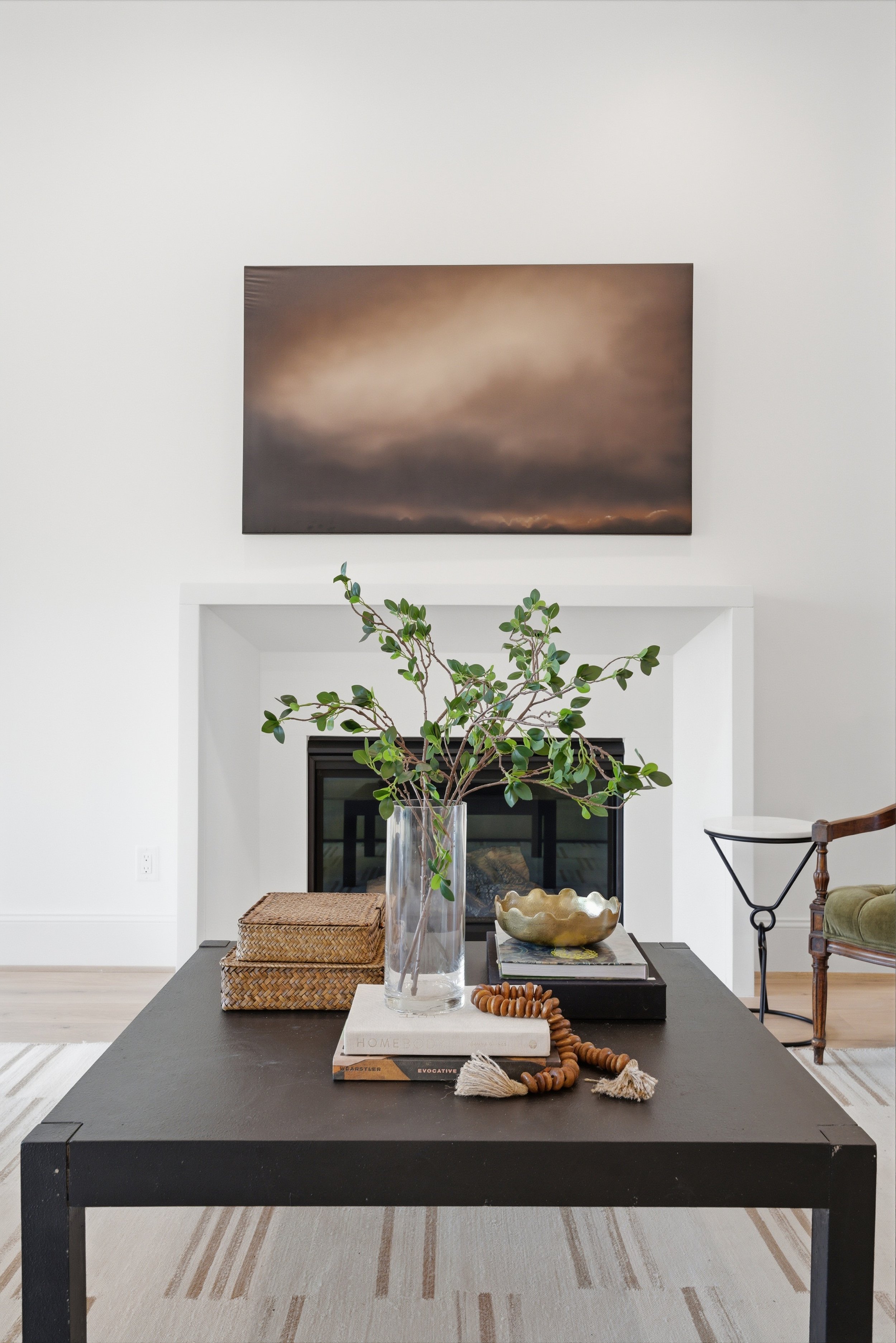 Living room with a black coffee table, a large glass vase with green branches, a woven basket, a gold decorative bowl, and books. A newspaper with a beaded necklace or bracelet on top, a fireplace with a framed artwork or photograph above, a side tab