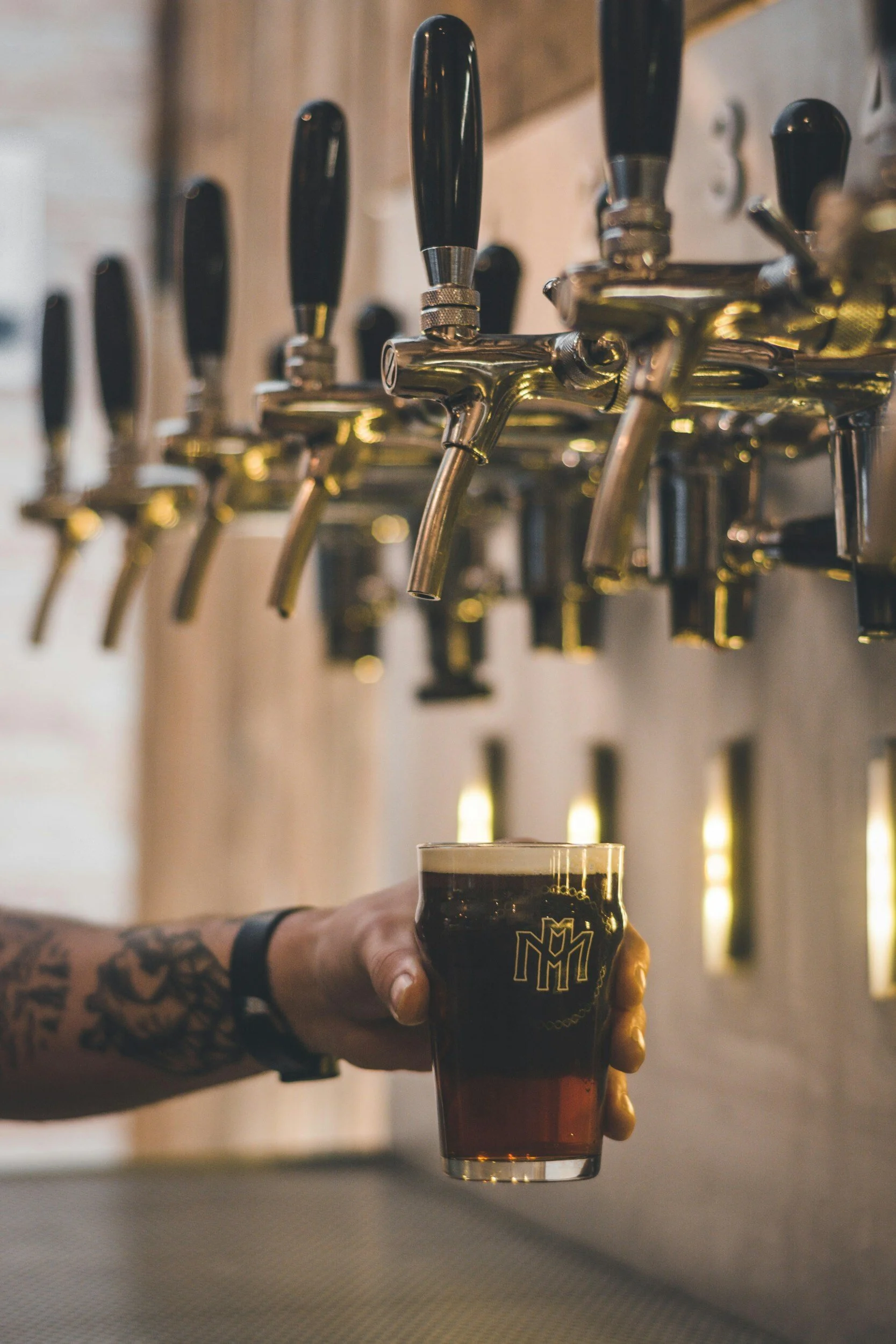 A person with tattoos on their arm is holding a glass of dark beer under multiple beer taps in a bar or brewery setting.