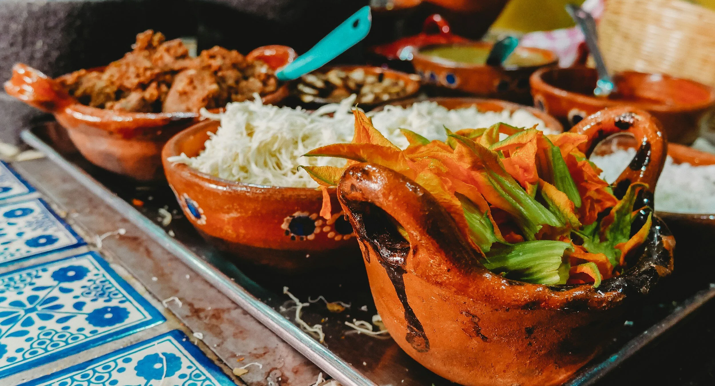 Traditional Mexican dishes in clay bowls, including shredded cabbage, fresh herbs, and cooked meat, on a metal tray with blue and white patterned tiles.
