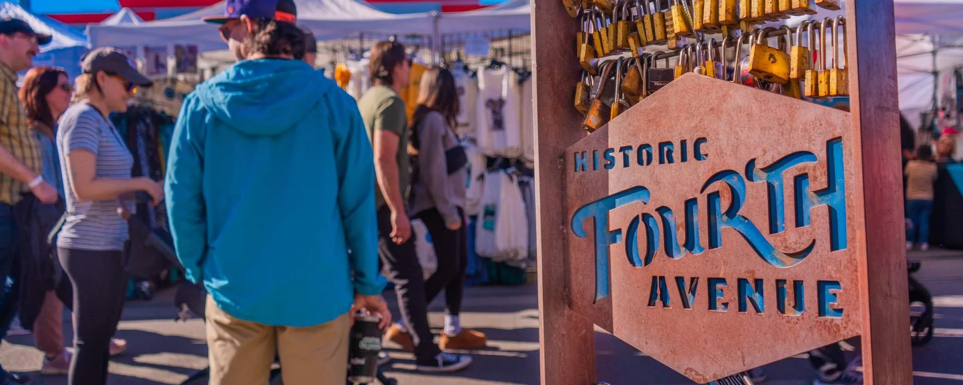 People walking along a street market with tents and stalls, and a sign reading "Historic Fourth Avenue" with padlocks attached to it.