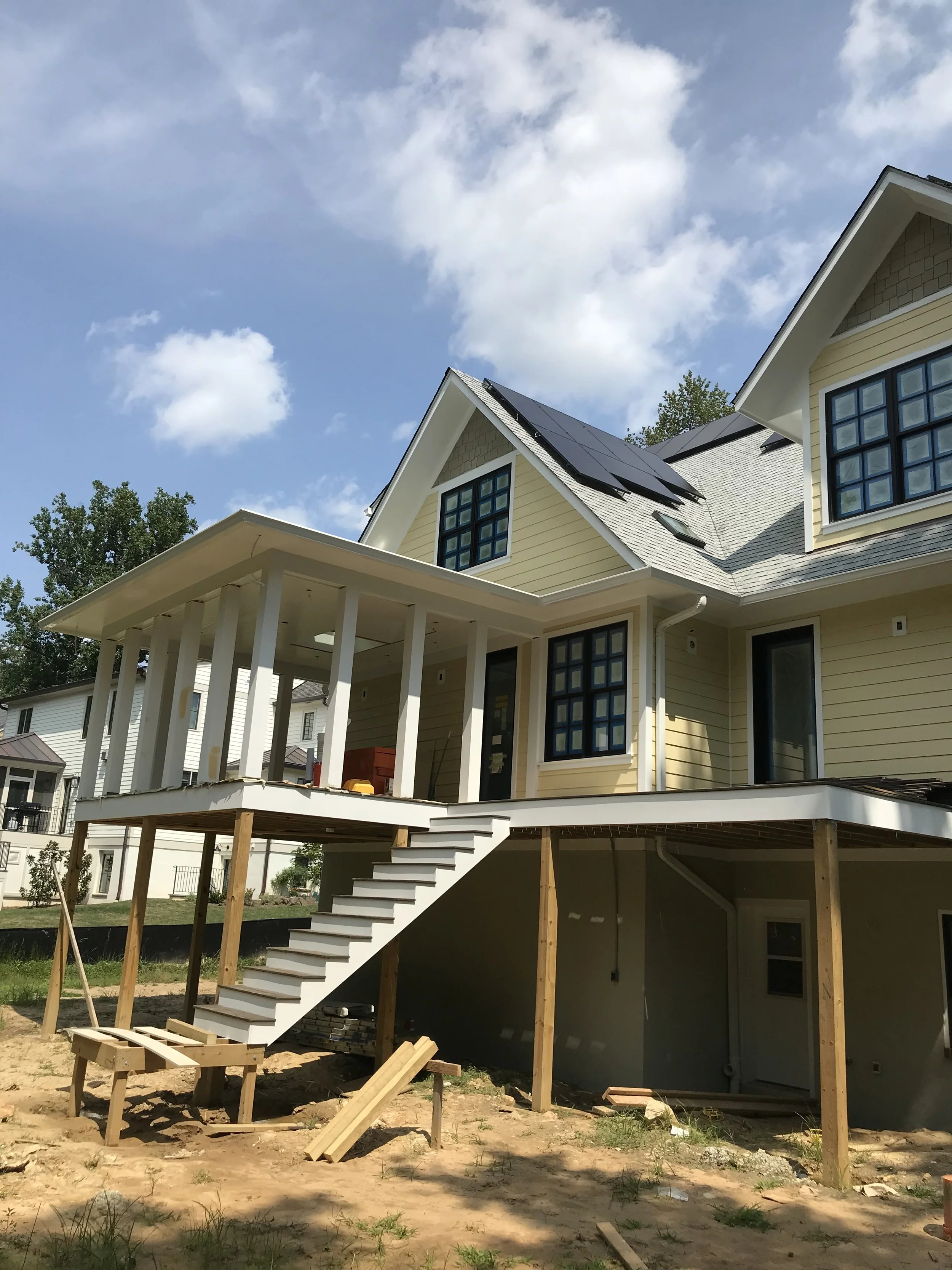 A yellow house under construction with an elevated porch, black solar panels on the roof, and a staircase leading up to the porch, in a neighborhood with other houses and a bright blue sky.