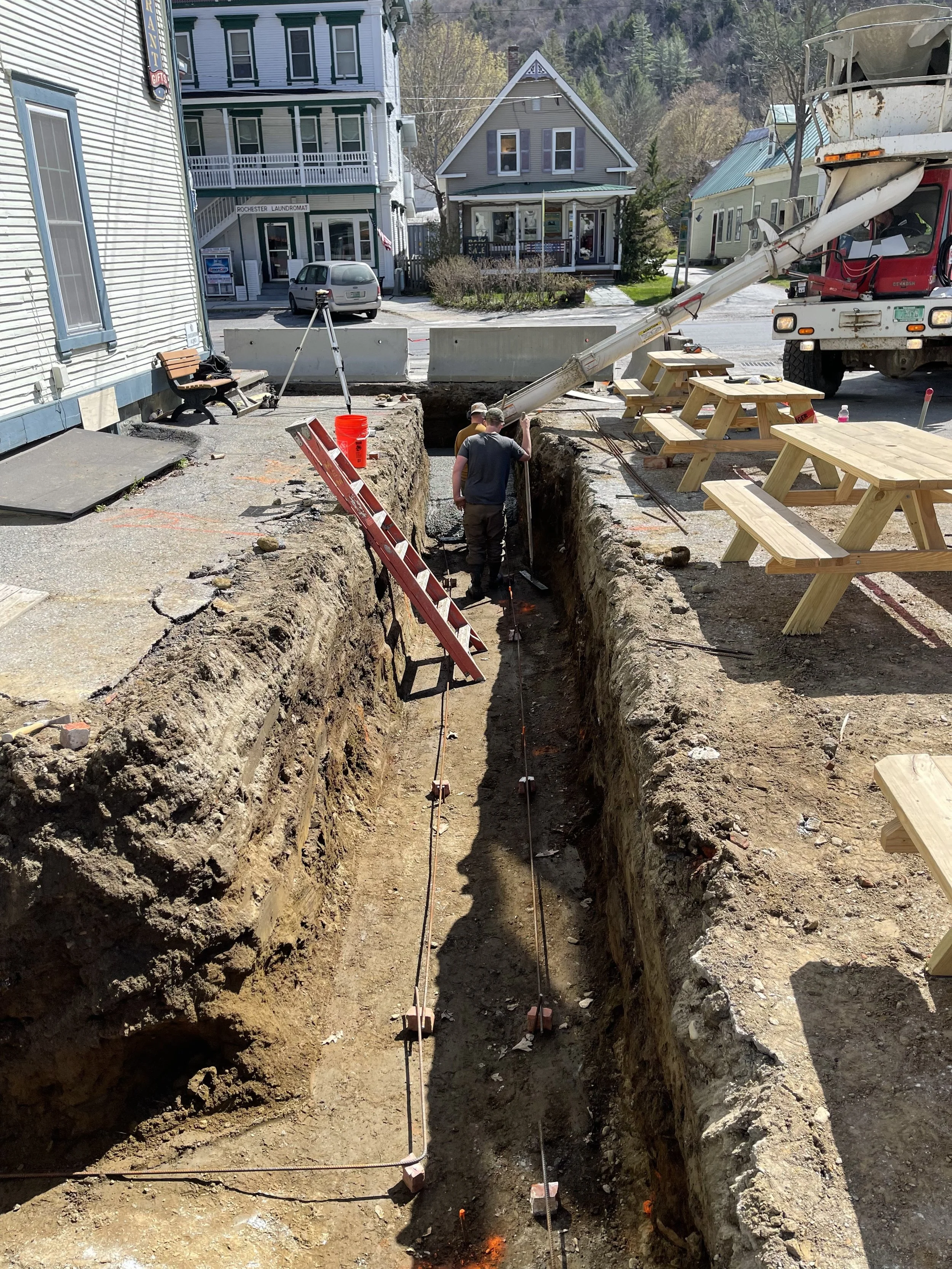 Construction workers underground in a trench with construction equipment and safety markers, in a small town with buildings, picnic tables, and a cement mixer truck nearby.