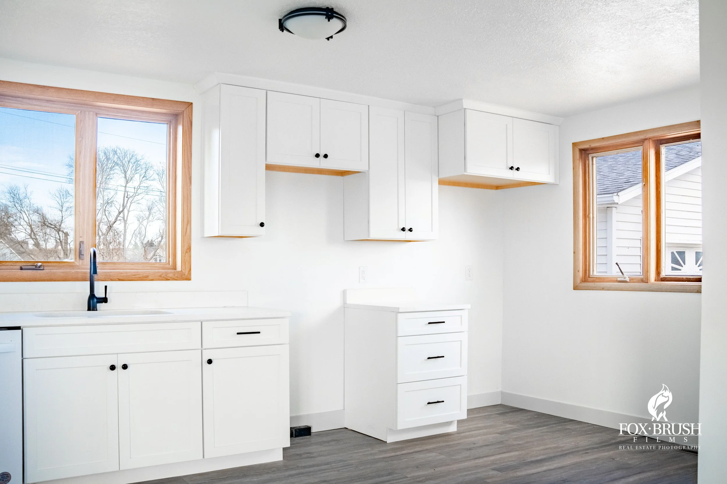 Empty kitchen with white cabinets, two windows with wooden frames, a black faucet, and light brown flooring.