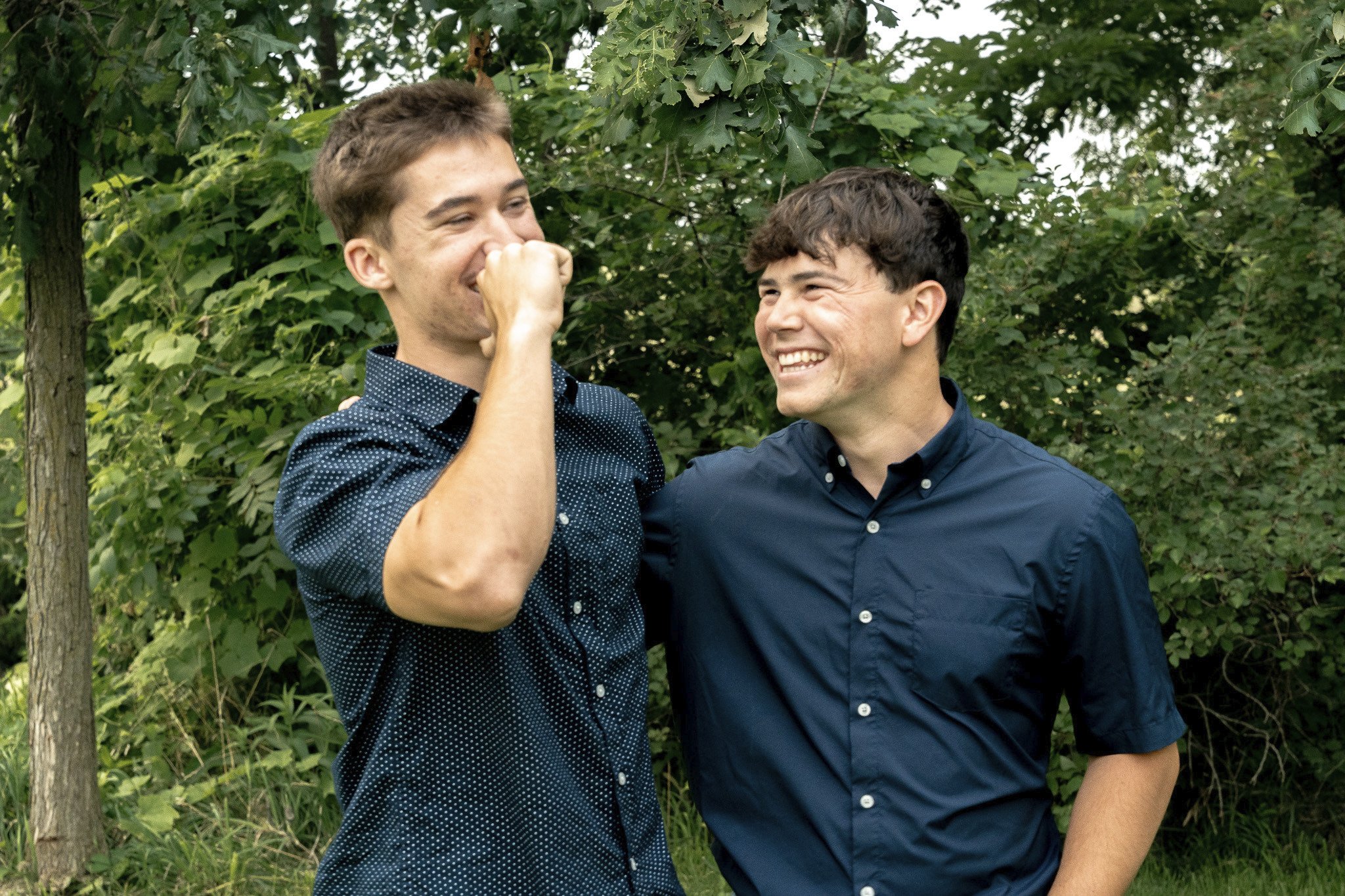 Two young men in blue shirts sharing a laugh in a green outdoor setting.