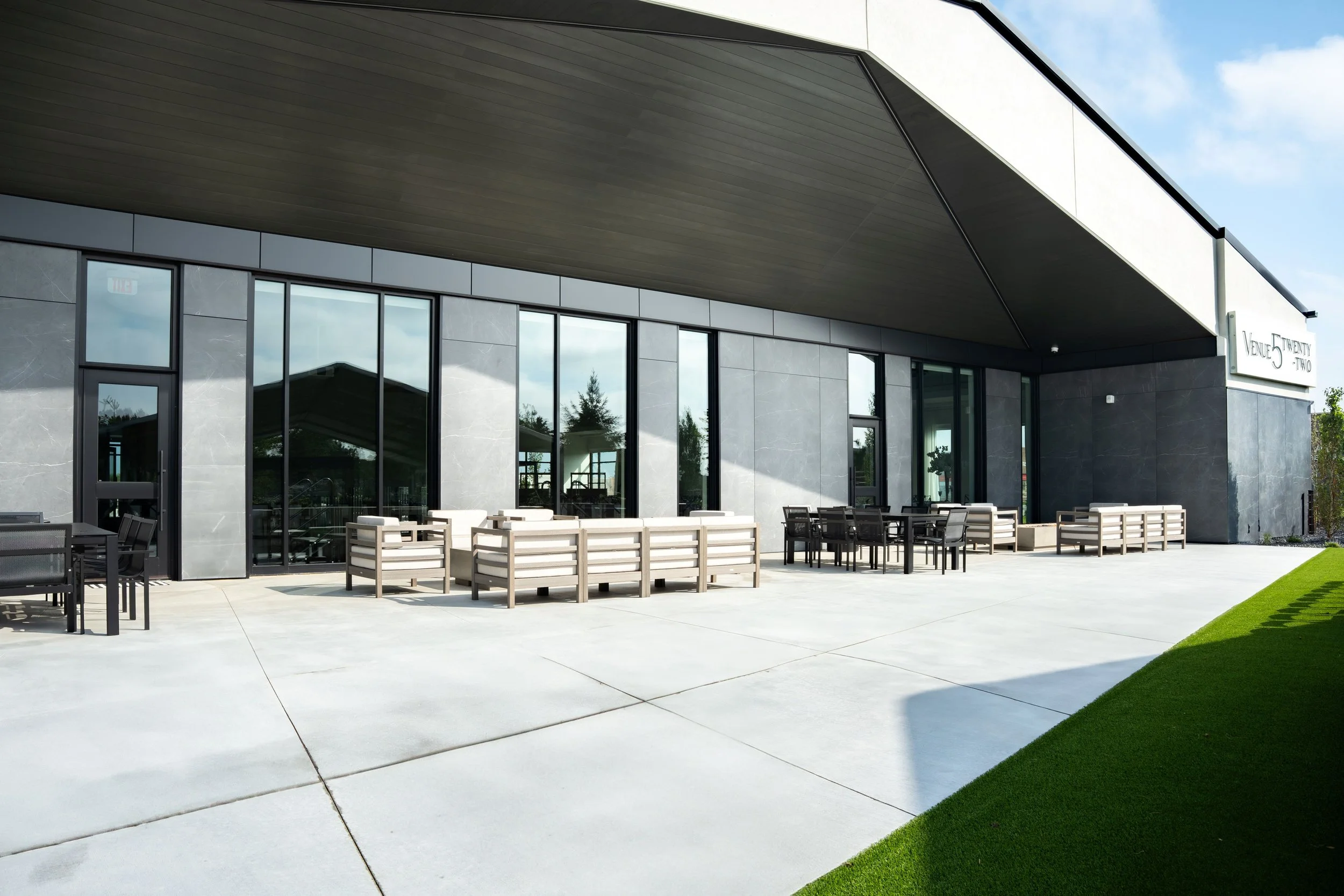 Modern building with large glass windows, outdoor seating with chairs and tables, concrete patio, and a grassy area with a slight incline.