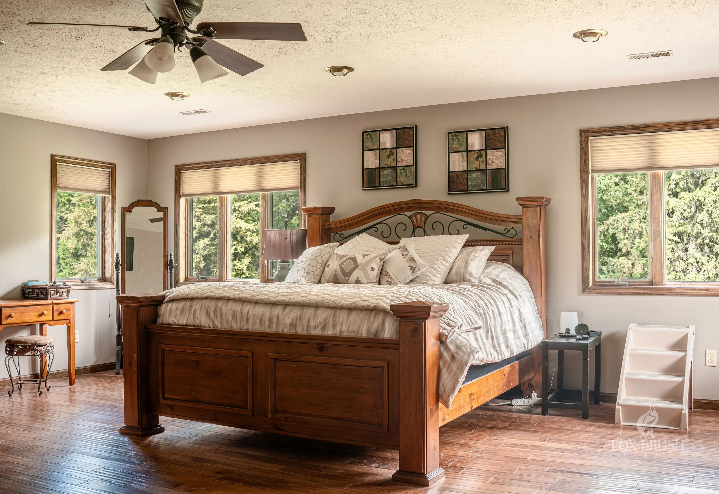 A bedroom with a large wooden bed, several pillows, and windows showing greenery outside. There's a ceiling fan, a nightstand with a lamp, a small black side table, and a white step stool.