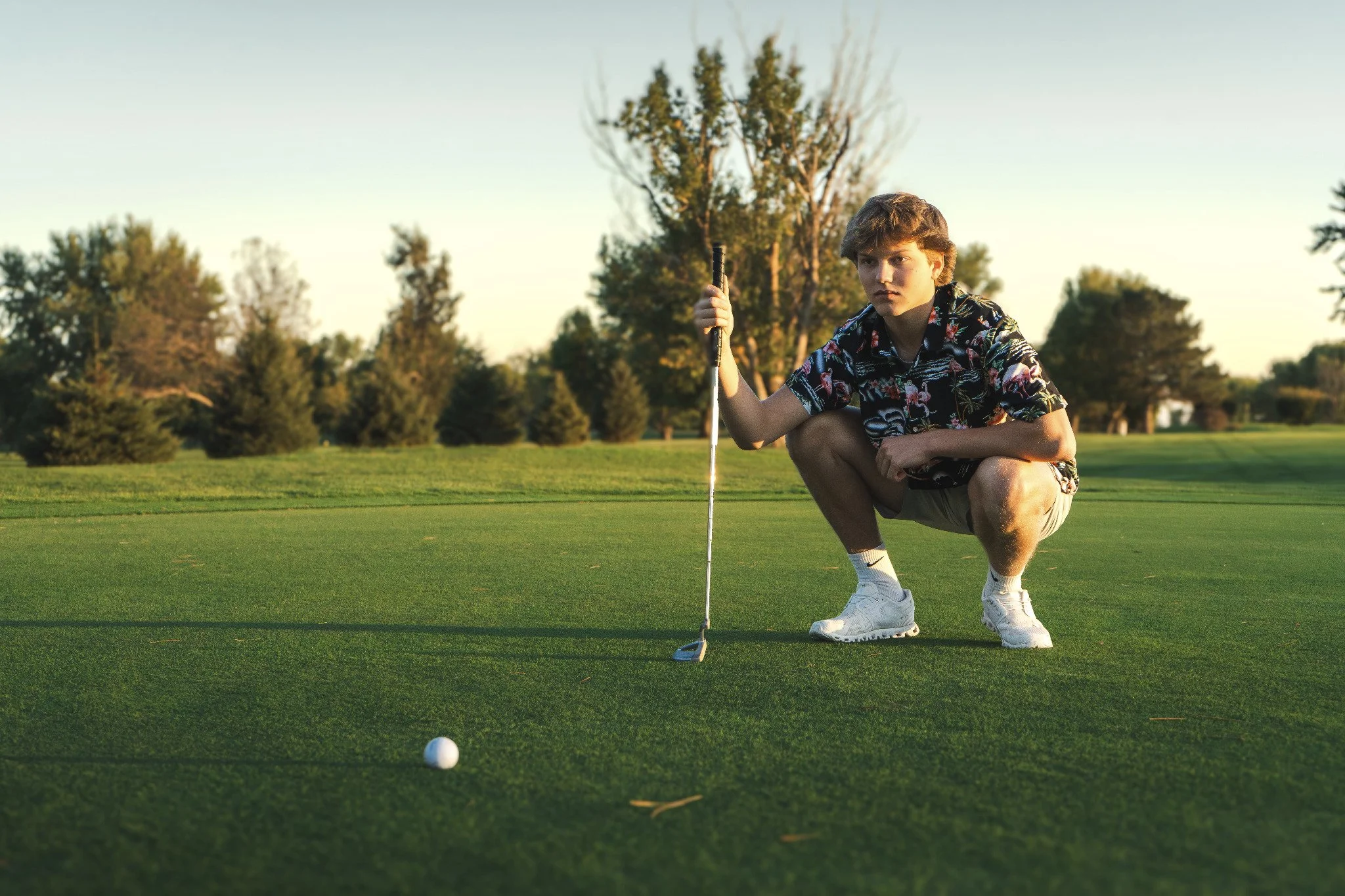 A young man crouching on a golf course, holding a golf club, preparing to putt a golf ball.
