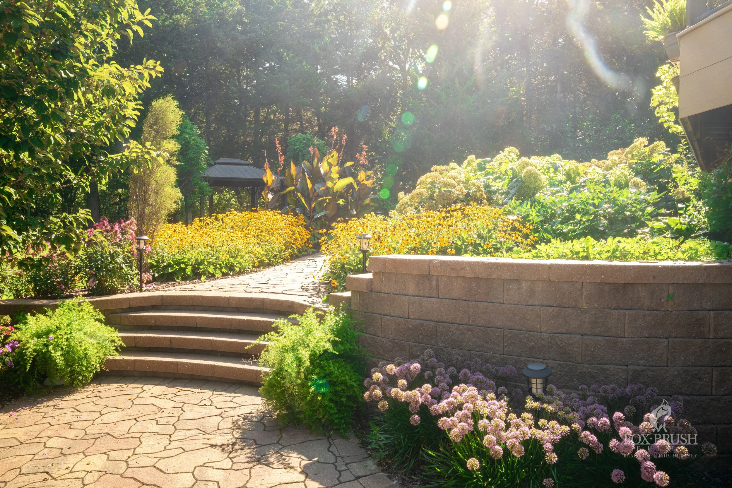 A landscaped garden with stone steps leading up to a pathway surrounded by lush flowering plants, bushes, and trees, with sunlight shining through the trees overhead.