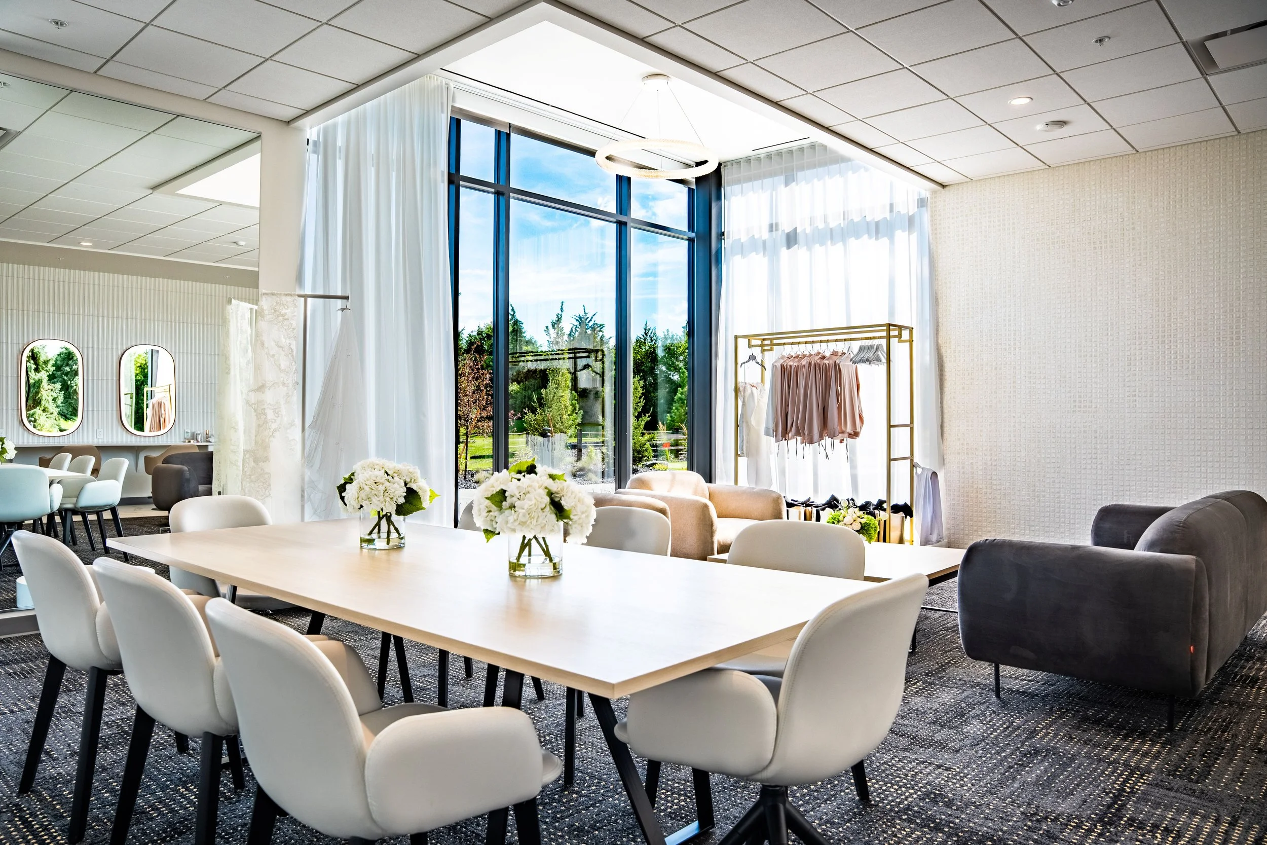 Bright, modern interior with large floor-to-ceiling windows, white curtains, a spacious white table with flower arrangements, and comfortable chairs and sofas, possibly a lounge or meeting space.