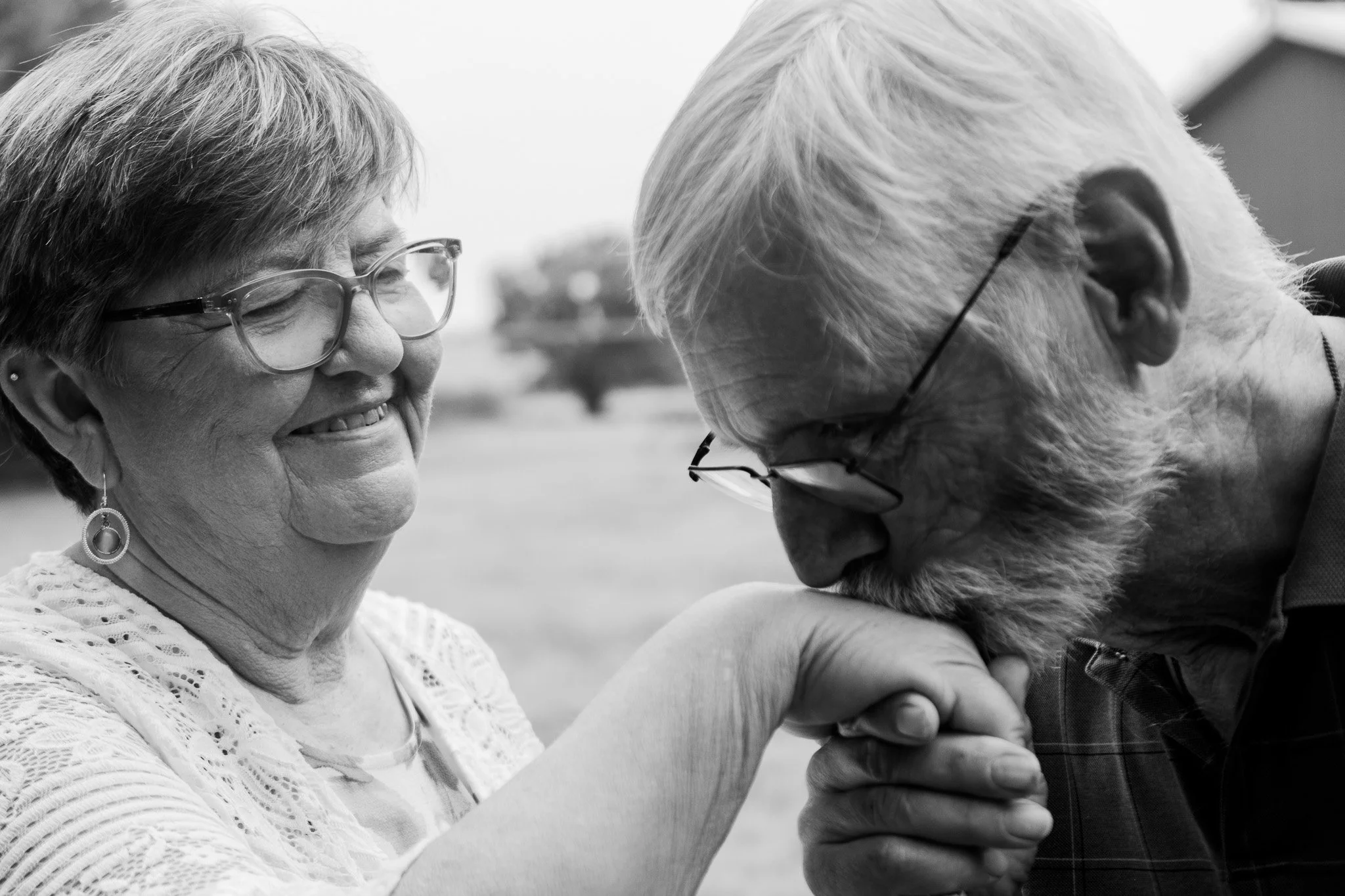 An elderly man kissing an elderly woman on the hand while she smiles. The woman wears glasses and earrings, and the man has glasses and a beard.