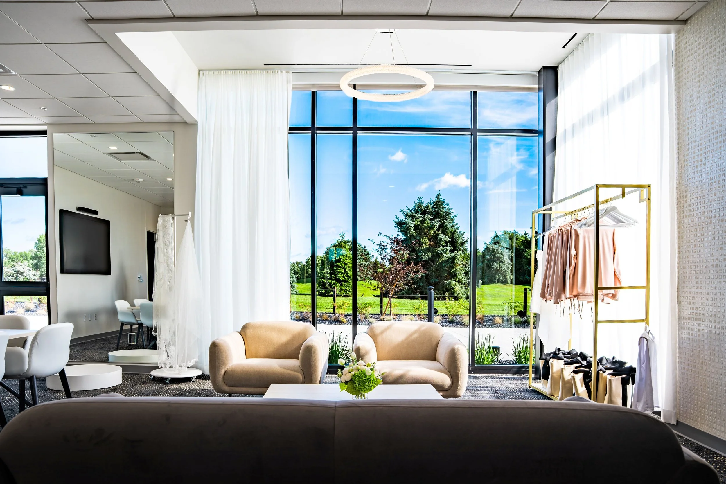 Modern lounge area with large floor-to-ceiling windows showing a green outdoor landscape, beige sofas, a white coffee table with a flower arrangement, white curtains, and a clothing rack with beige and white garments.