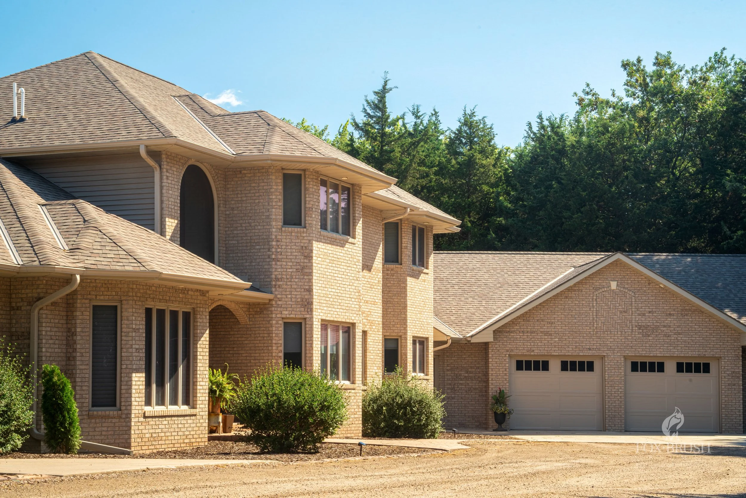 A large two-story brick house with multiple windows, a sloped roof, and a attached garage with two doors. There are green bushes and a potted plant near the house, with a dirt driveway in front and a wooded area in the background.