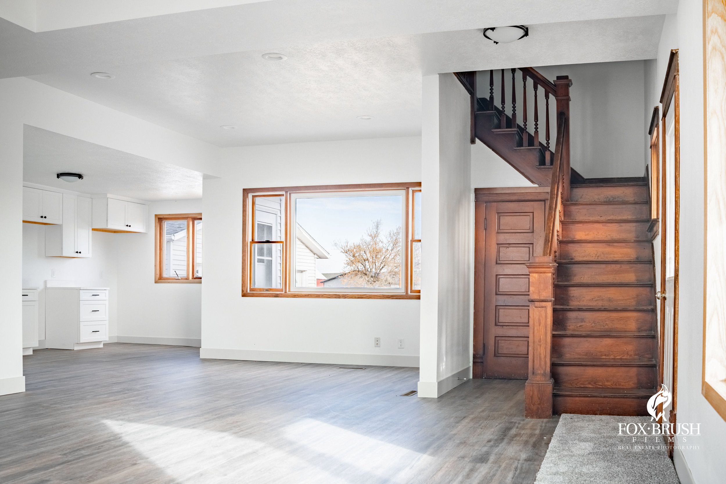Empty living room with large window, white walls, gray wood flooring, and a wooden staircase.