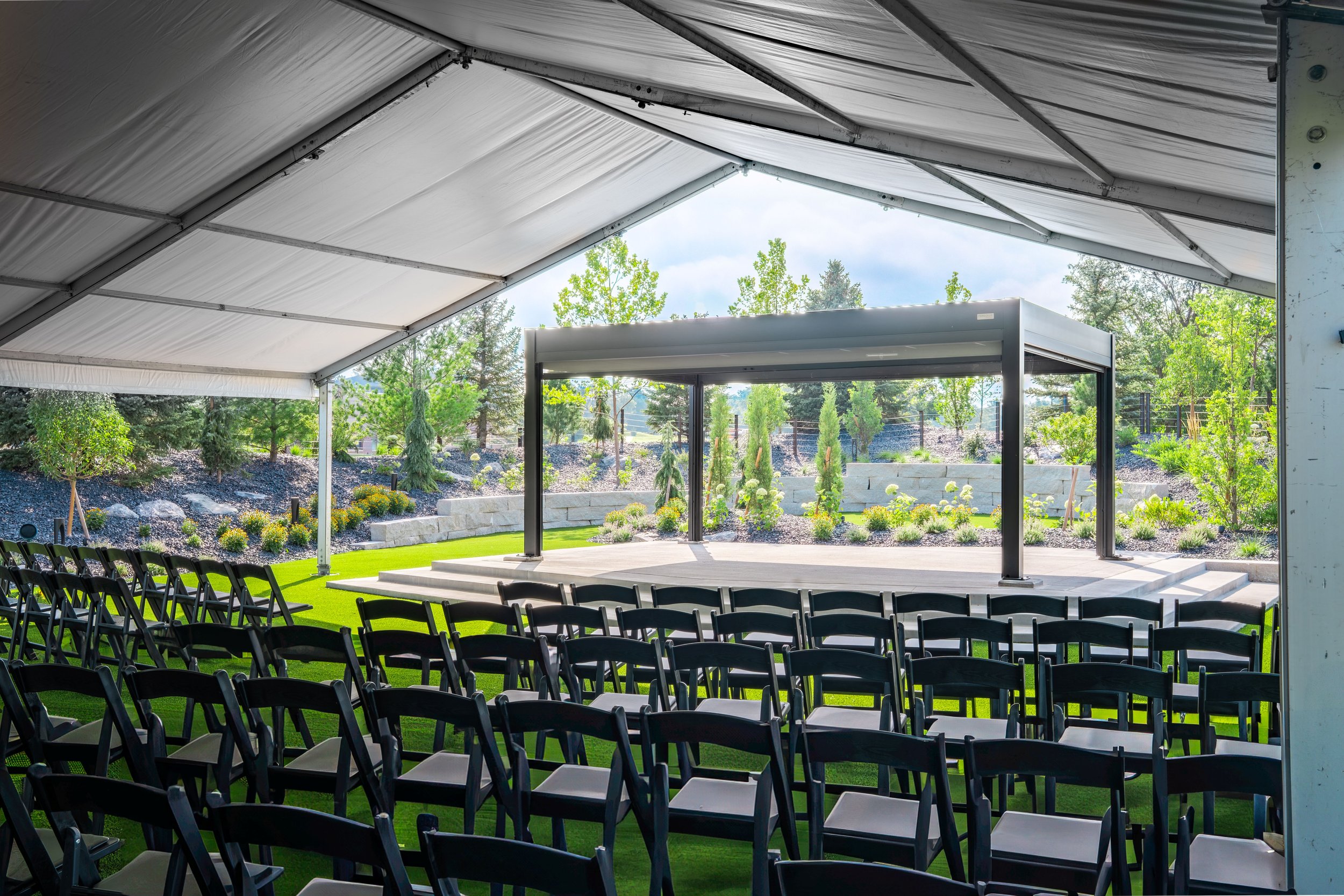 Outdoor event space with rows of black chairs, a small stage, and a canopy roof with garden and trees in the background.