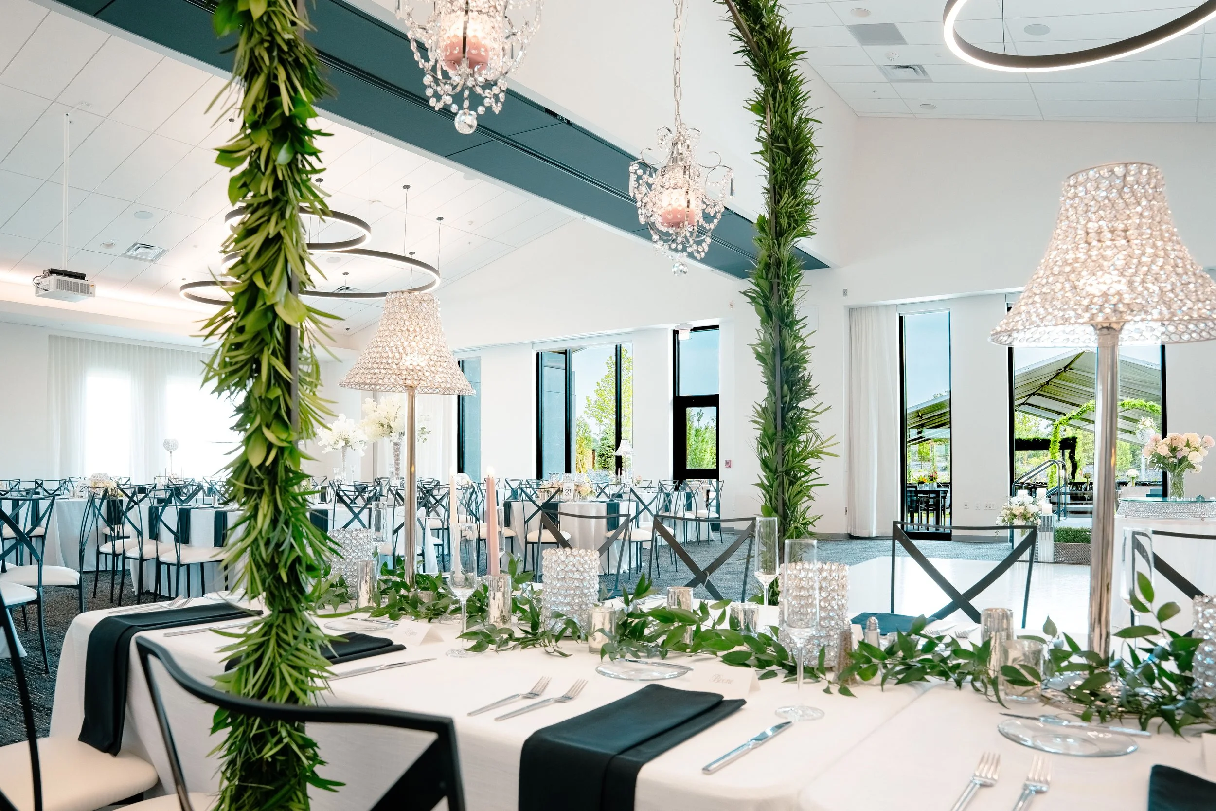 Elegant banquet hall decorated for a wedding reception with white and black color scheme, green foliage accents, large windows, and chandeliers.