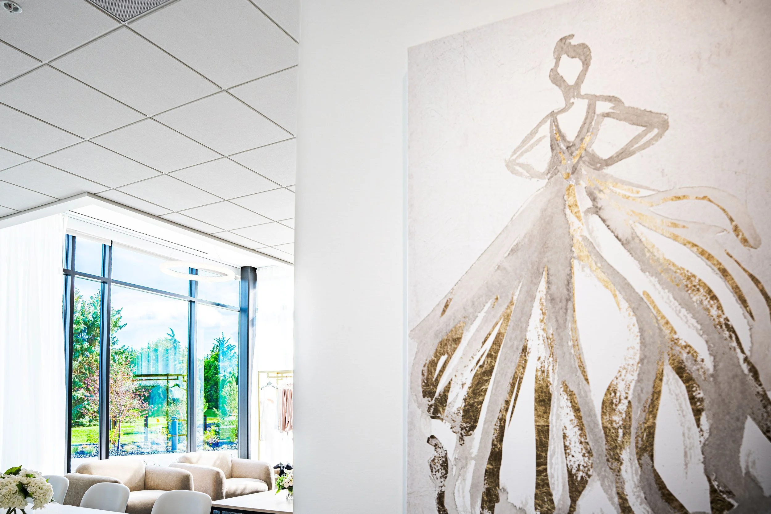 Interior of a bright, modern room with large glass windows showing trees outside. There is a beige sofa and white chairs with flower arrangements on the tables. A large wall features a stylish, abstract painting of a woman in a flowing dress.