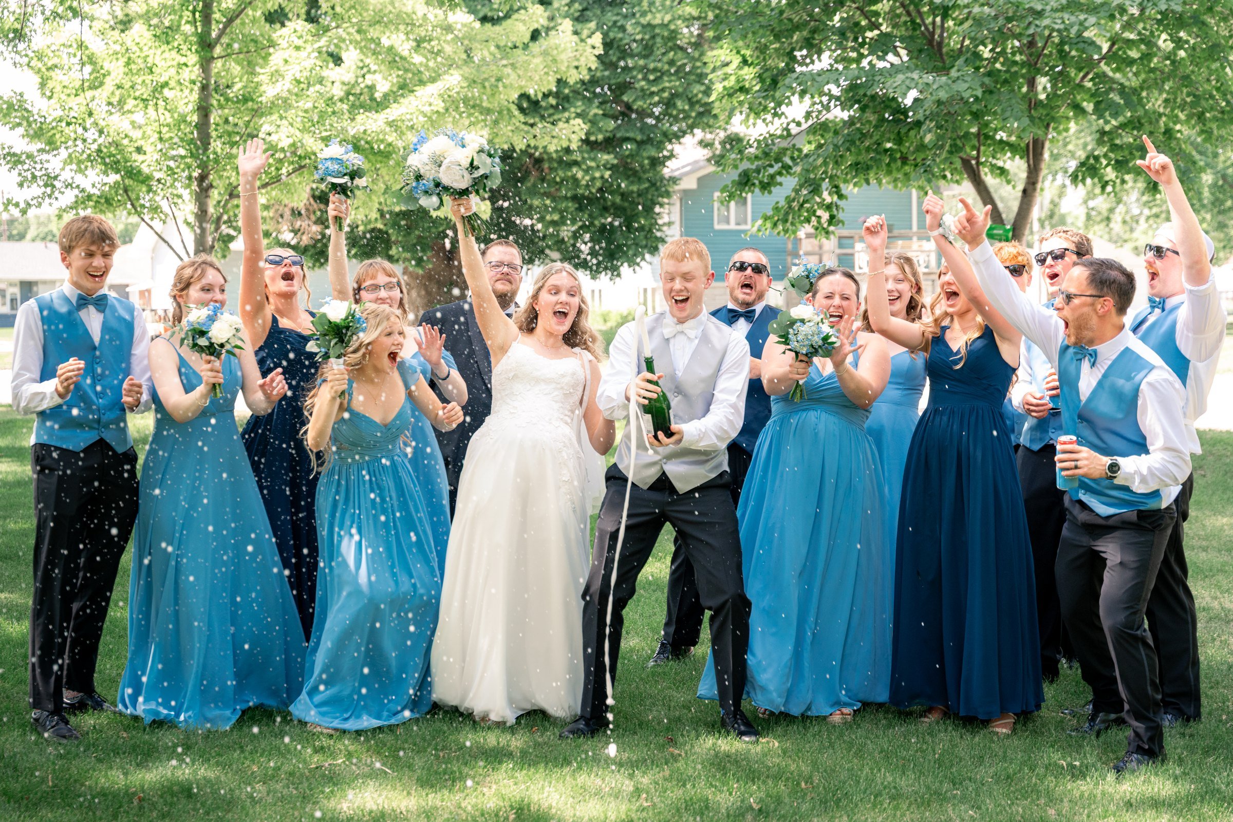 Wedding party cheering outdoors around the bride and groom, with bridesmaids in blue dresses.