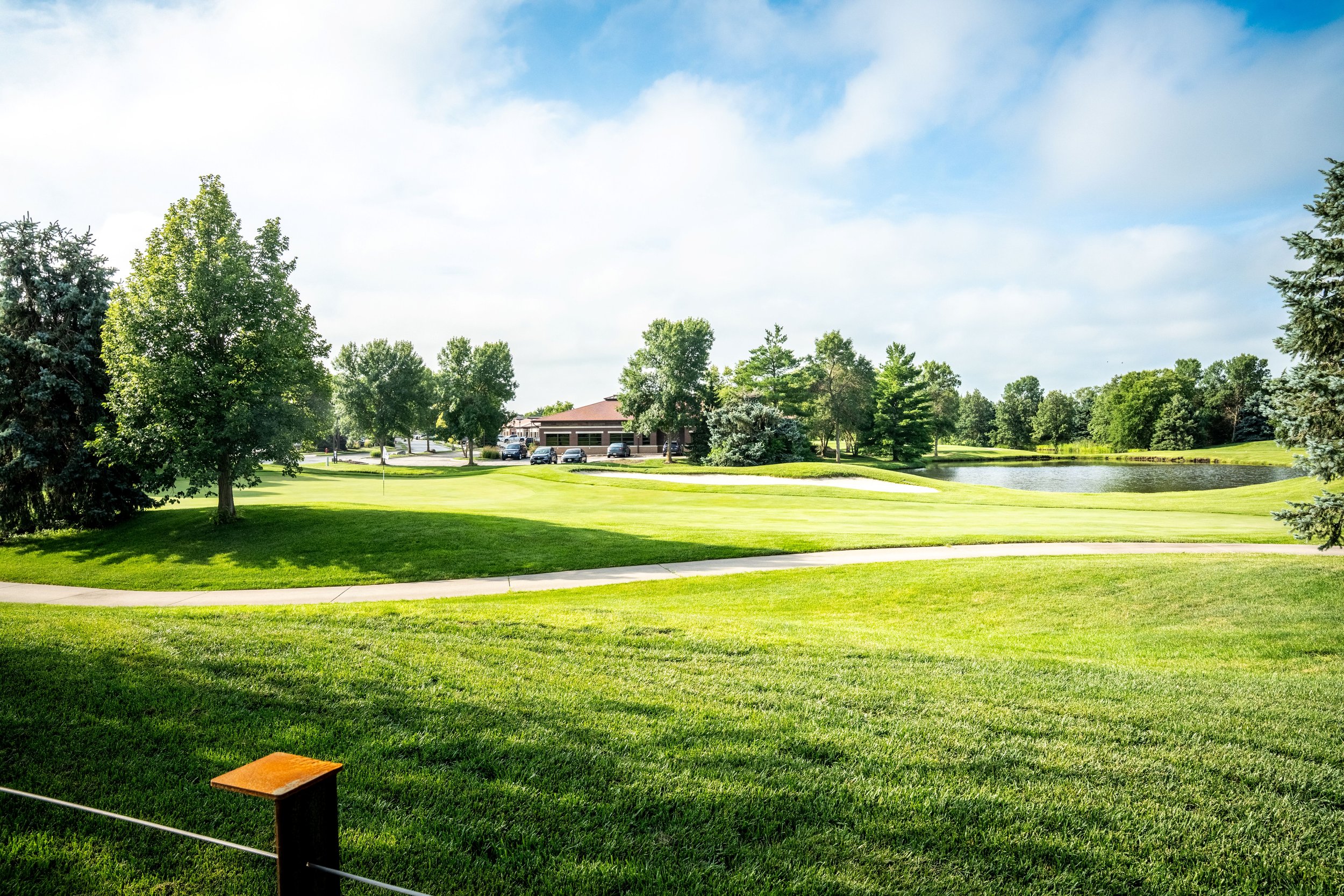 A golf course with well-maintained green grass, trees, a small pond, and a building in the background under a partly cloudy sky.