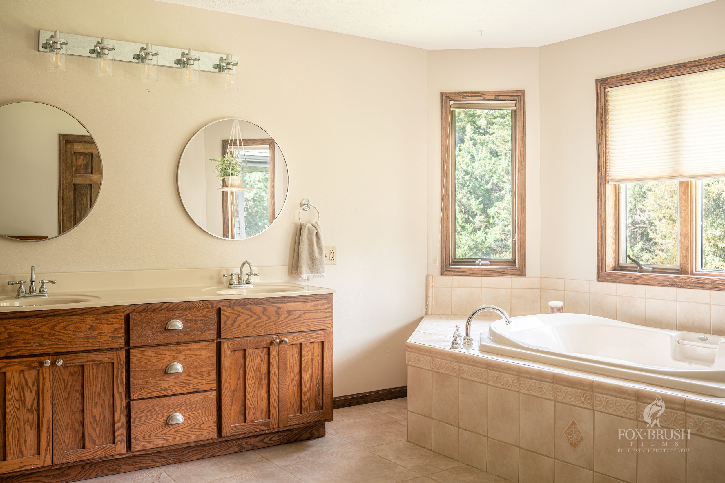 Bathroom with double sinks, wooden cabinets, two round mirrors, natural light, tub with windows, tiled floor.
