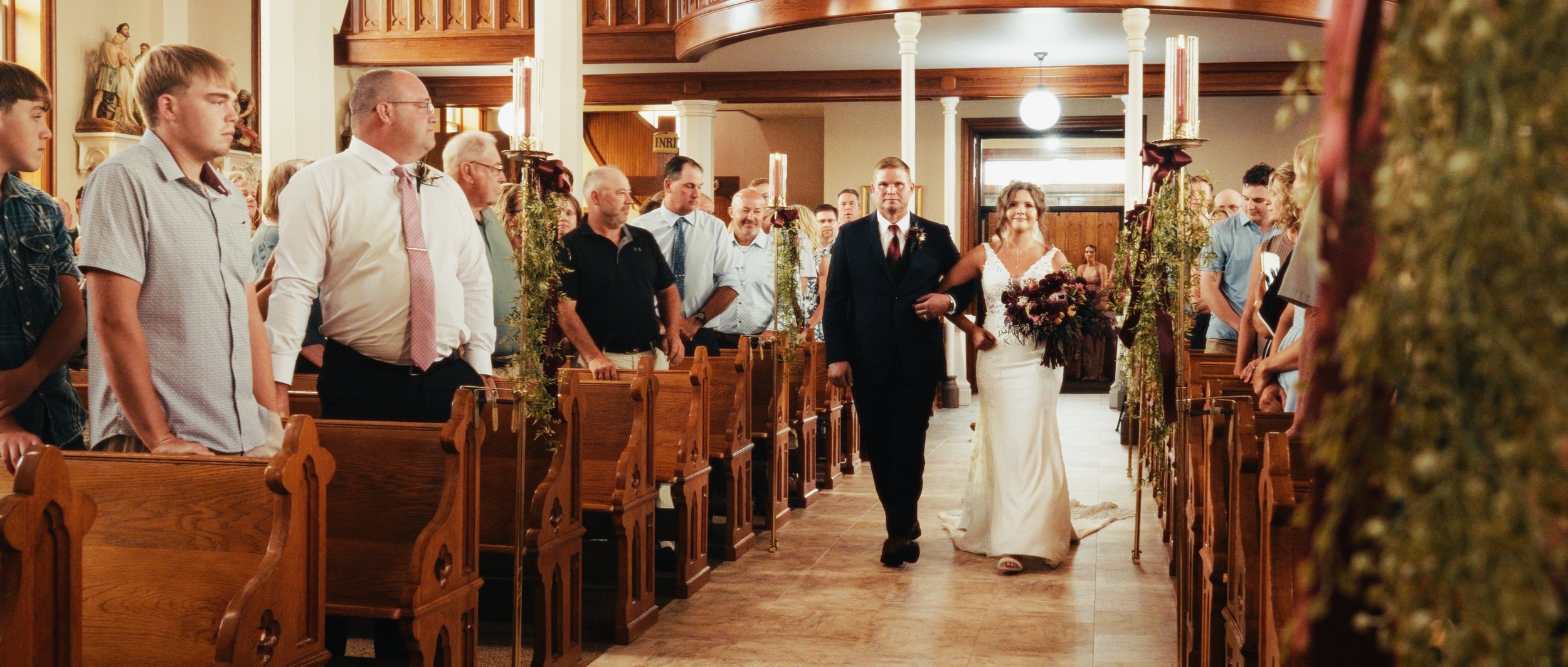 A bride in a white wedding dress walking down the aisle with a man in a dark suit during a wedding ceremony in a church filled with guests.