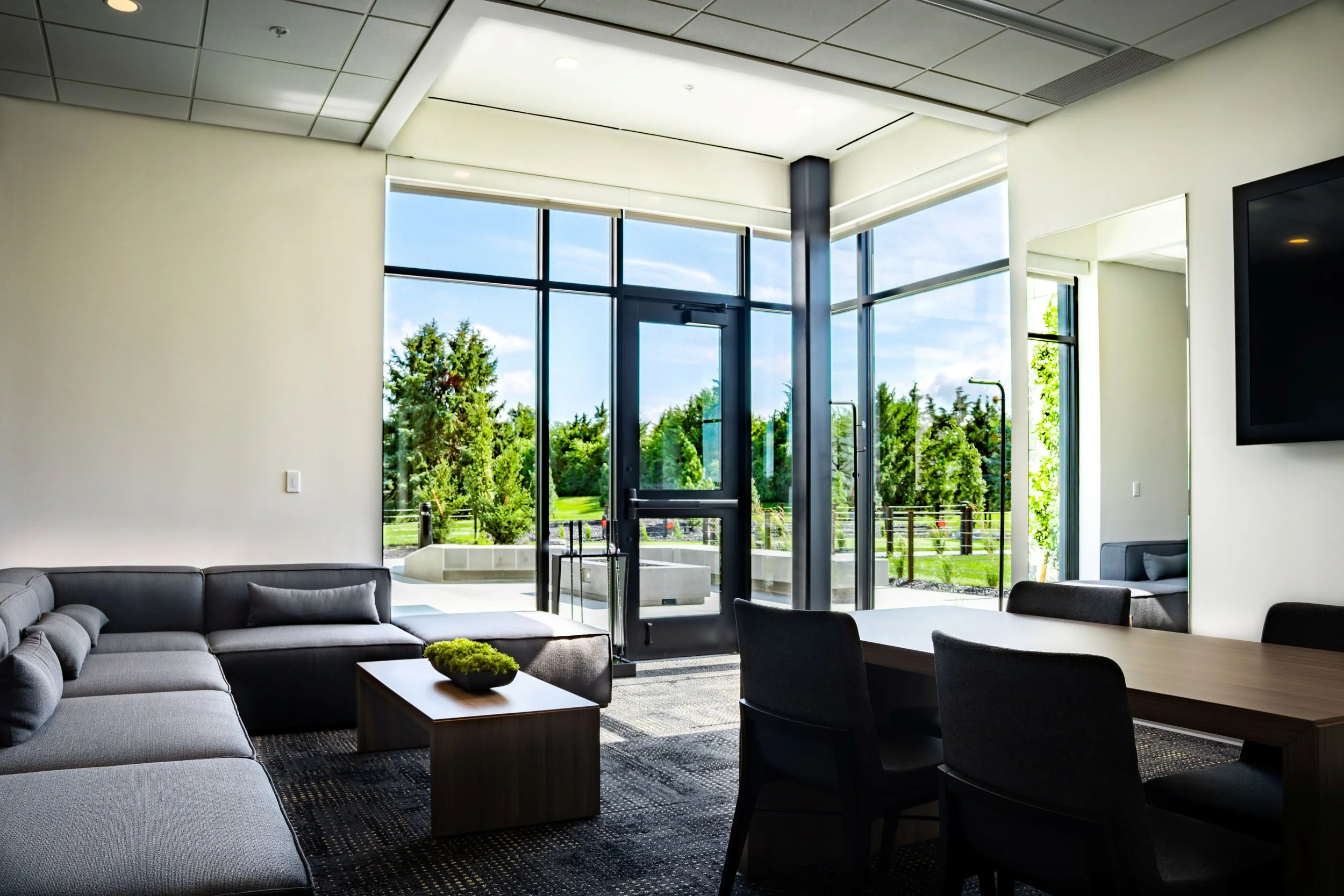 Modern lounge area with large glass windows showing a green outdoor space, gray sectional sofa, and a wooden dining table with black chairs.