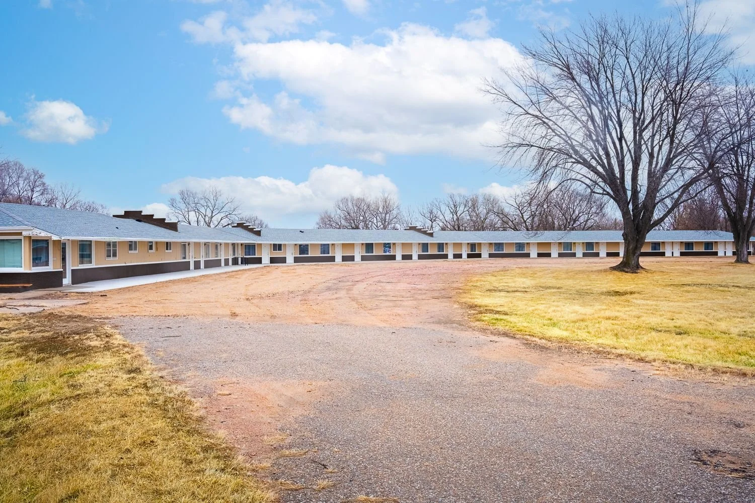 A row of single-story beige motel rooms with white trim and blue roofs, surrounded by a grassy lawn, large leafless trees, and a partly cloudy sky.