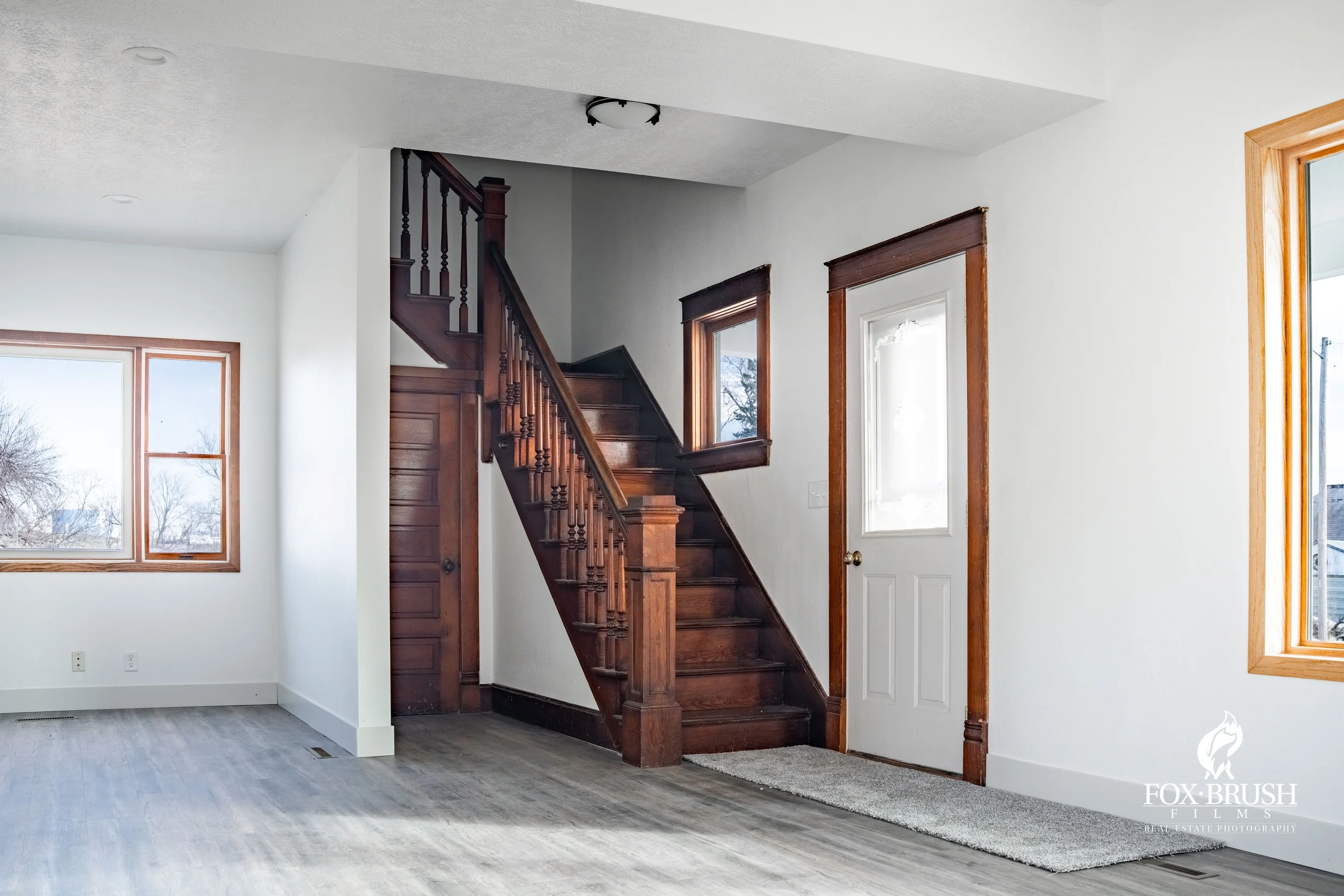 Living room with wooden staircase, white walls, large windows with wooden trim, and a white door with a window panel.