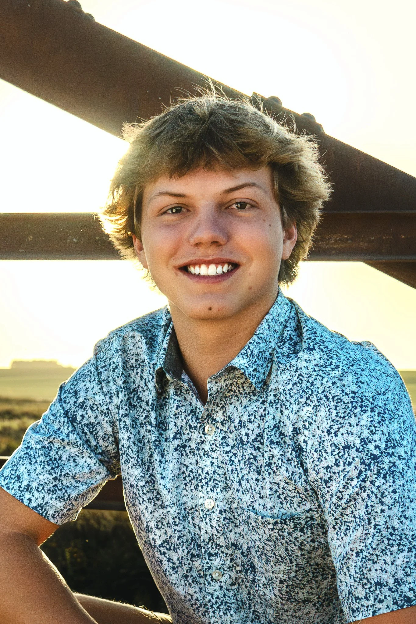 A young man with tousled light brown hair smiling at the camera, wearing a patterned short-sleeve shirt, with outdoor scenery and a metal bridge structure in the background during sunset.