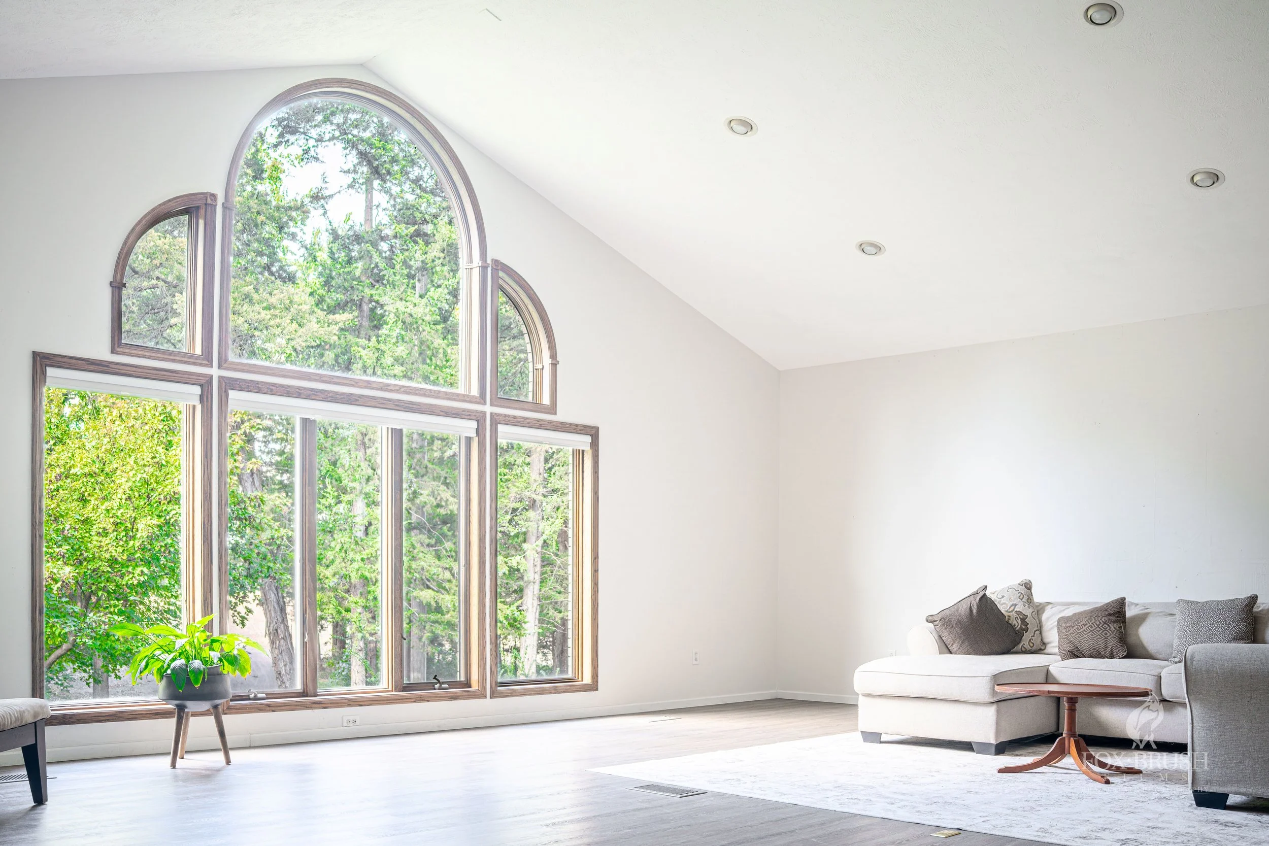 Living room with large arched windows showing green trees outside, white walls, a white sectional sofa with gray and patterned pillows, a small wooden side table, a potted plant, and recessed ceiling lights.