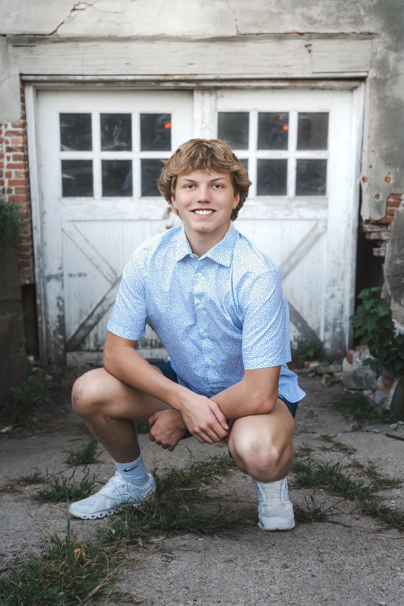 A young man with curly brown hair, wearing a light blue polka-dot shirt, shorts, and white athletic shoes, squatting on a concrete surface outdoors in front of an old, weathered white garage door and brick wall.