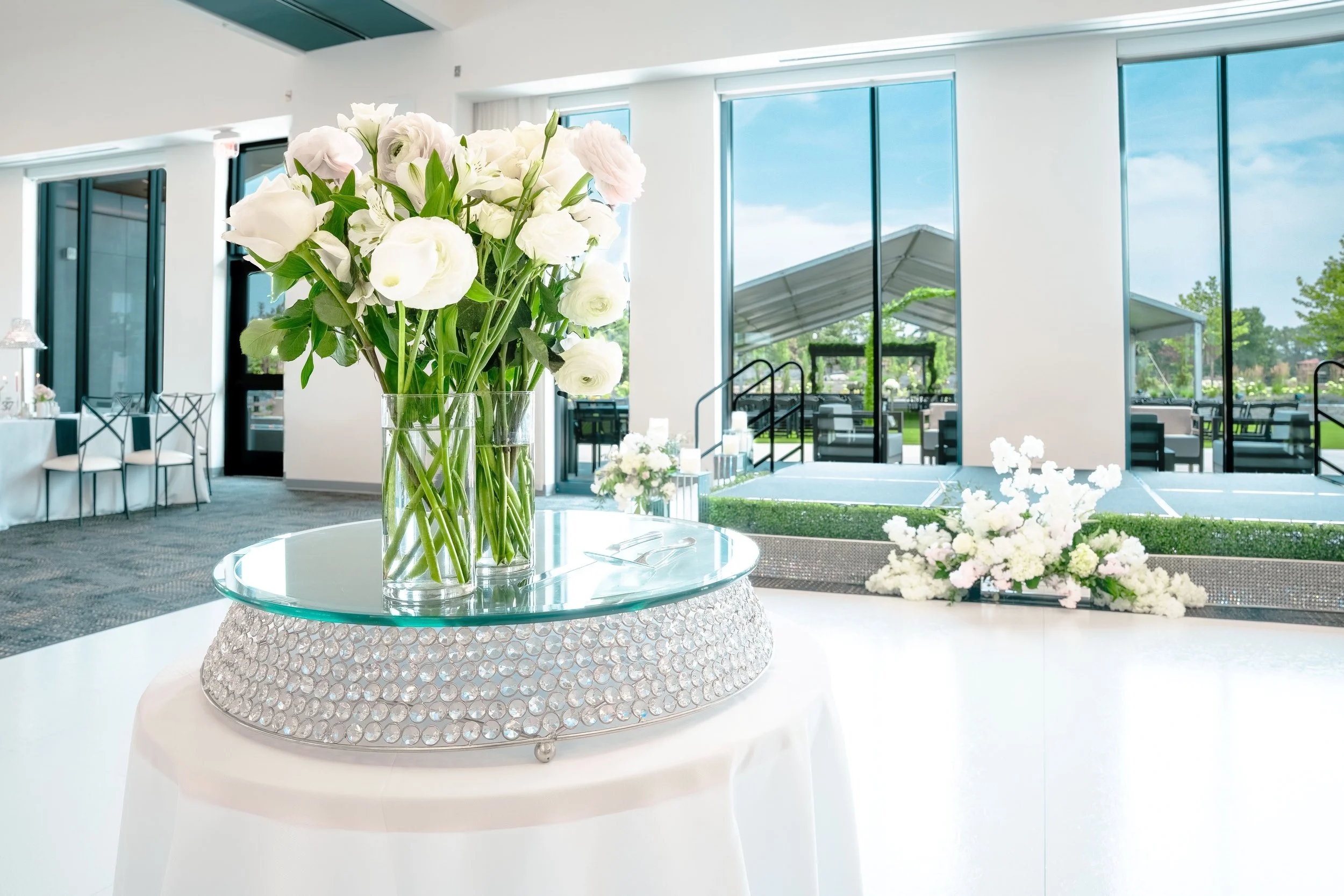 Elegant event space with floral centerpieces, large glass windows with views of outdoor tables and greenery, and a raised stage area.
