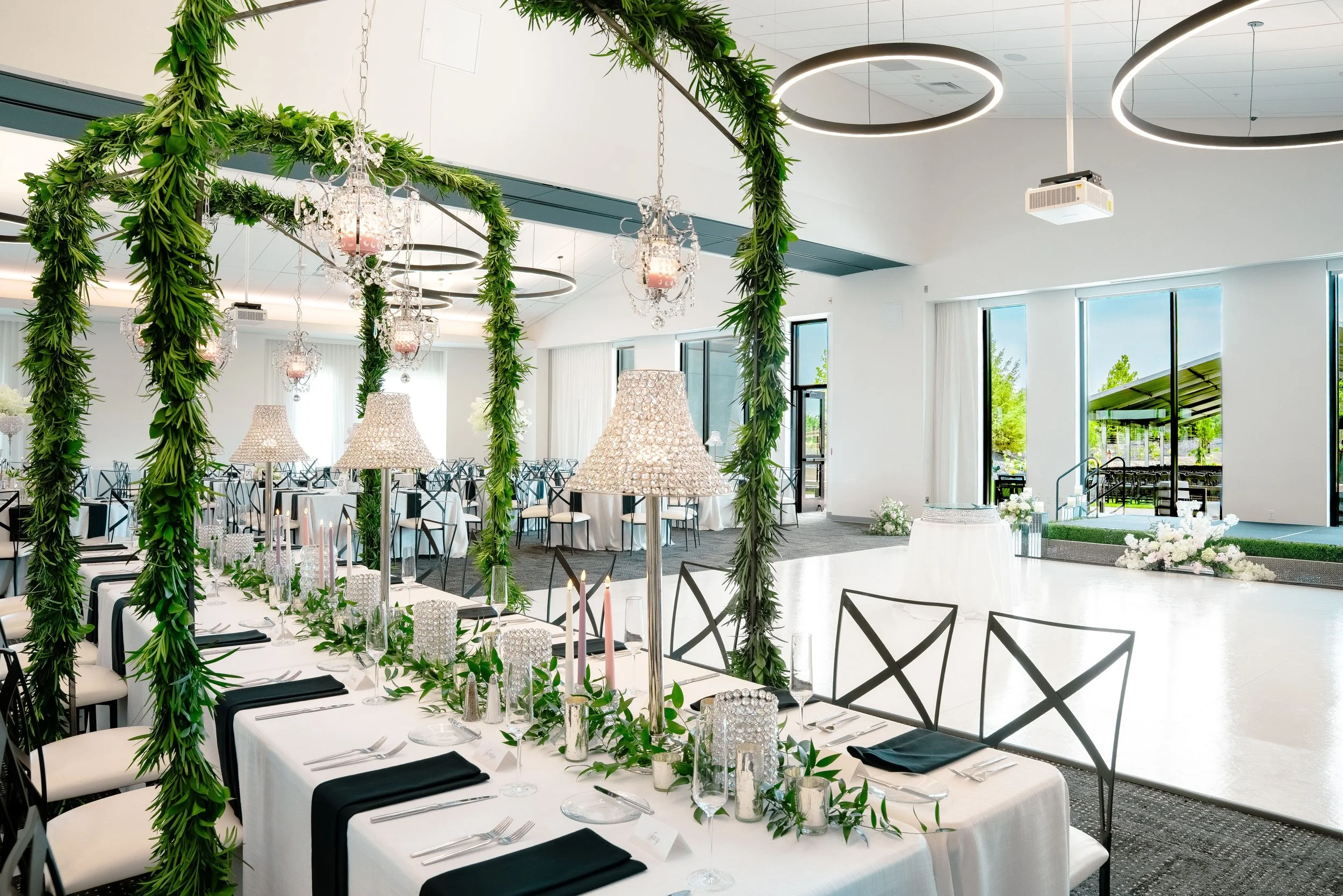 Elegant indoor wedding reception with decorated tables, greenery arches, and large windows letting in natural light.