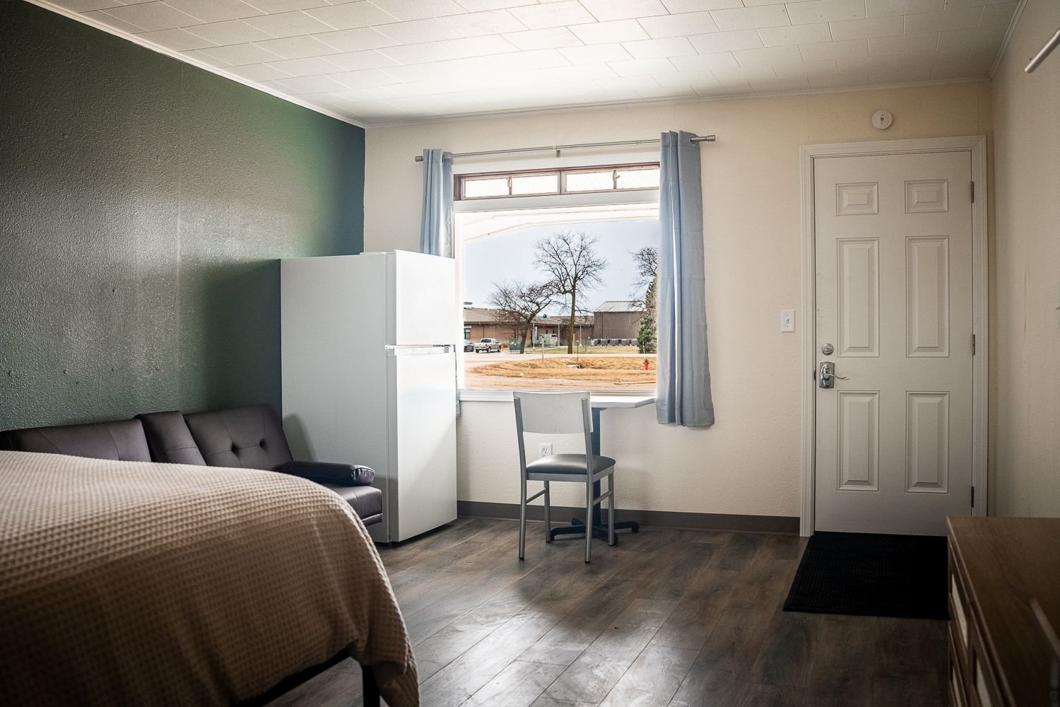 An interior of a simple studio apartment room with a bed, a black sofa, a white refrigerator, a chair, a window with blue curtains, and a door, with wooden flooring and beige walls.