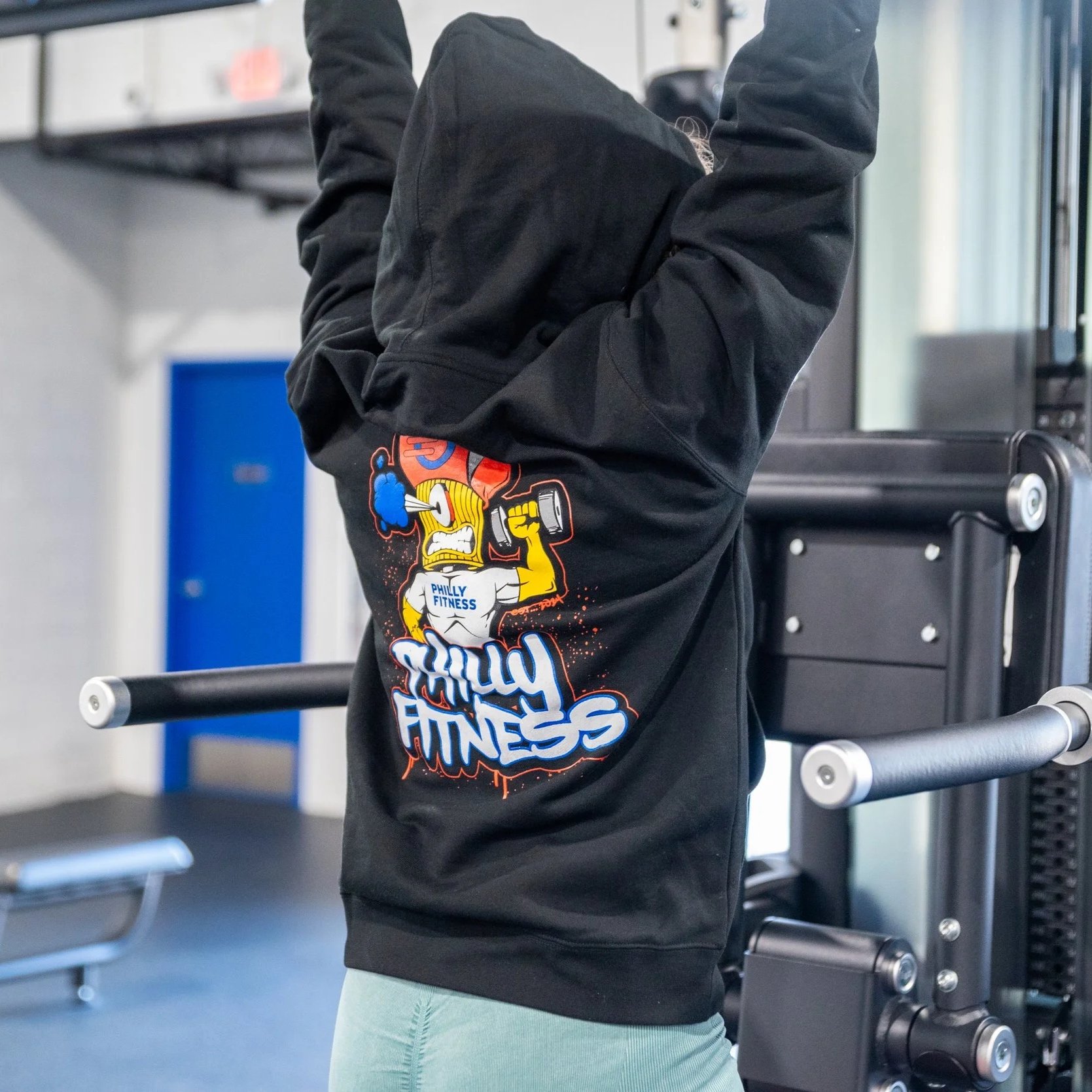 Gym member using the pull up bar while wearing the Philly Fitness® Graffiti Graphic Hoodie on a side profile