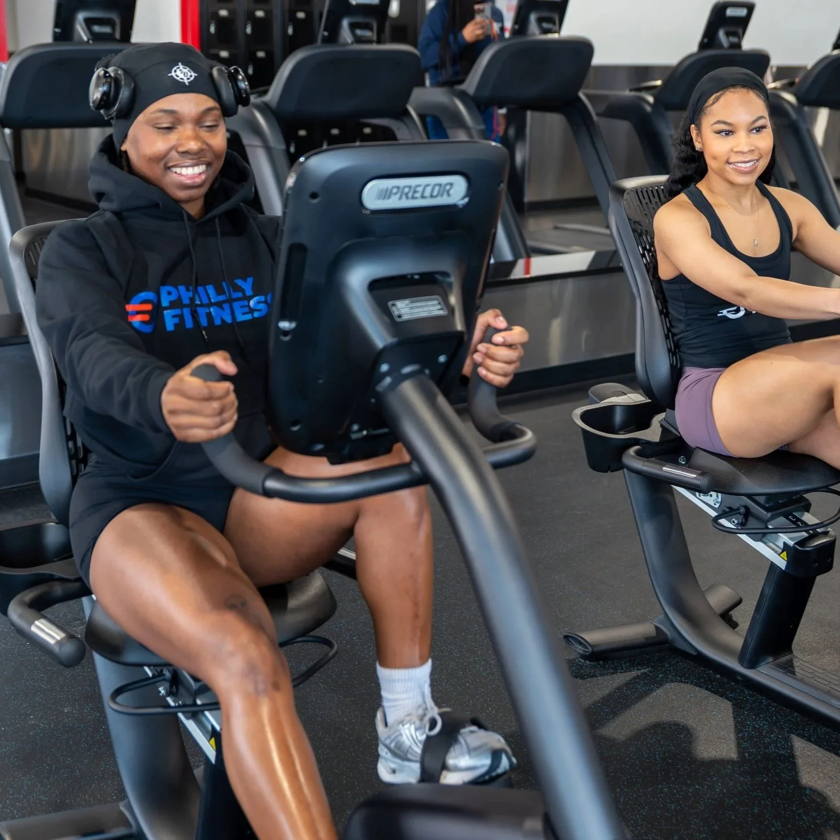 Gym members on the bike in the PhillyFitness® Black Hoodie with Full Color Logo