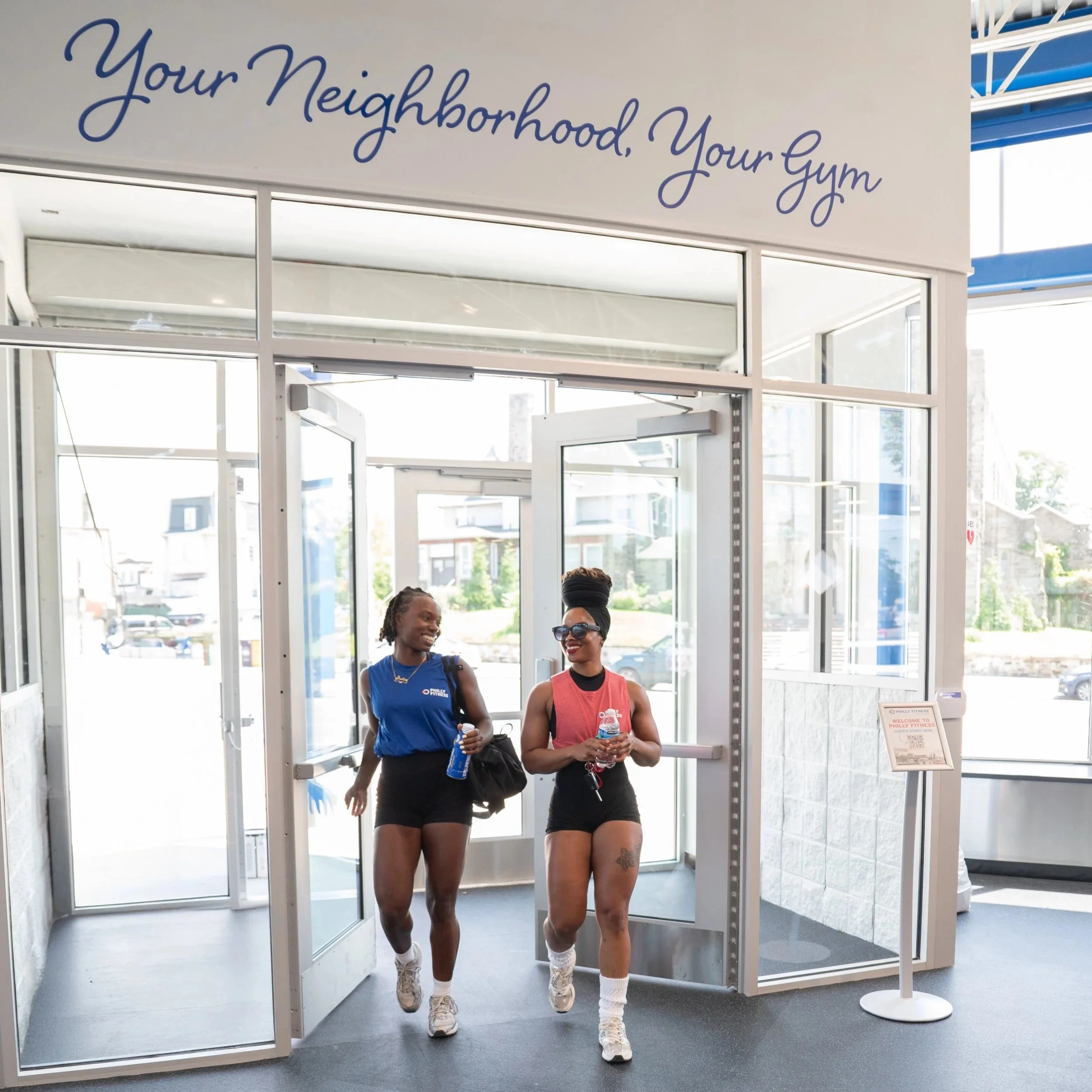 Two gym members walking into Philly Fitness® wearing a Philly Fitness® Blue Mens and Red Womens Muscle T-Shirt