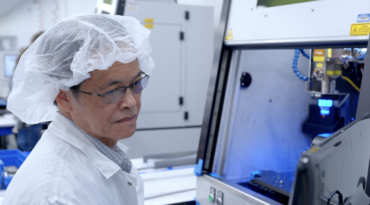 Cirtec Medical employee in a hairnet and lab coat standing beside a manufacturing machine inside a cleanroom