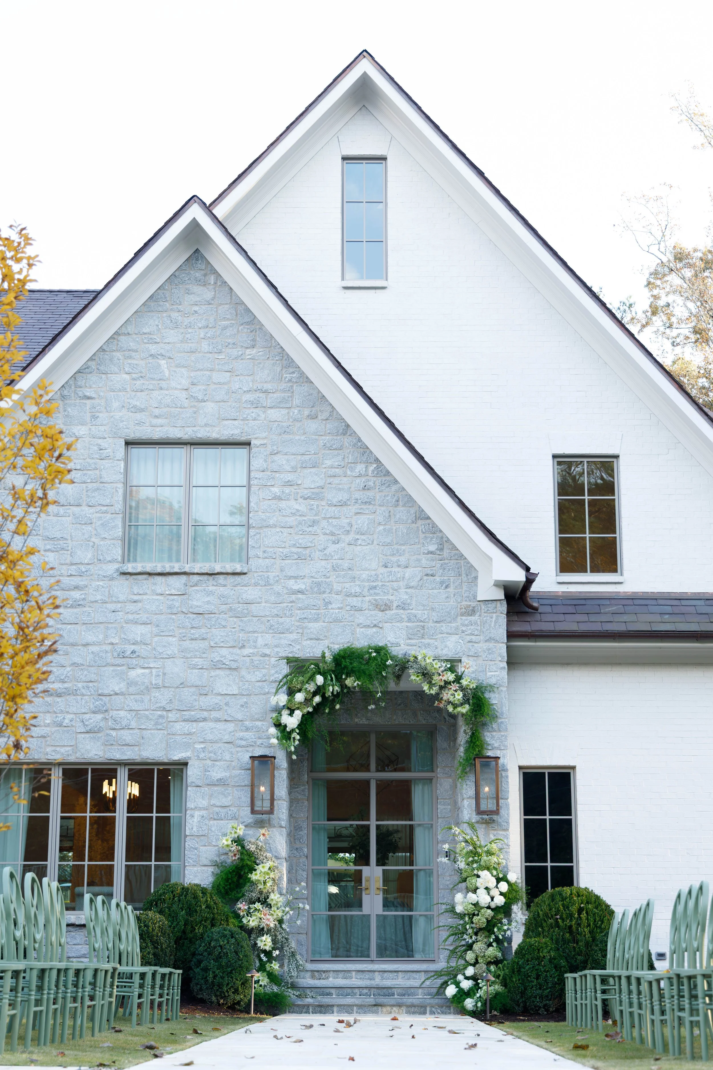 Front view of a white house with gray stone accents, decorated with flowers and greenery for a wedding or special event, featuring glass doors, windows, and green chairs along a paved walkway.