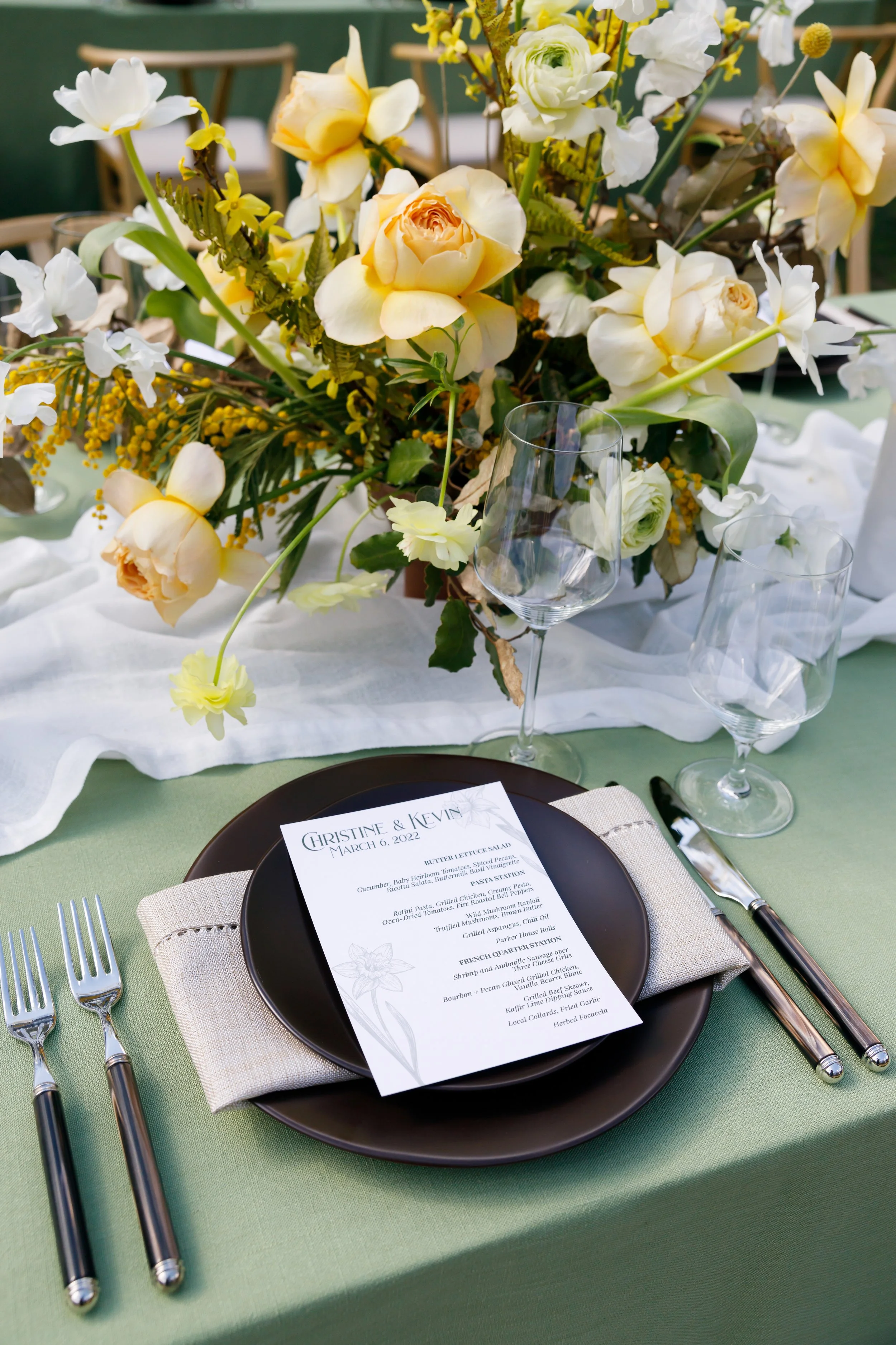 Elegant table setting with a dark plate, beige napkin, silverware, and a printed menu for Christin & Kevin's wedding on March 6, 2022. Decorative floral centerpiece with yellow and white flowers on a white table runner, two empty wine glasses, and a 