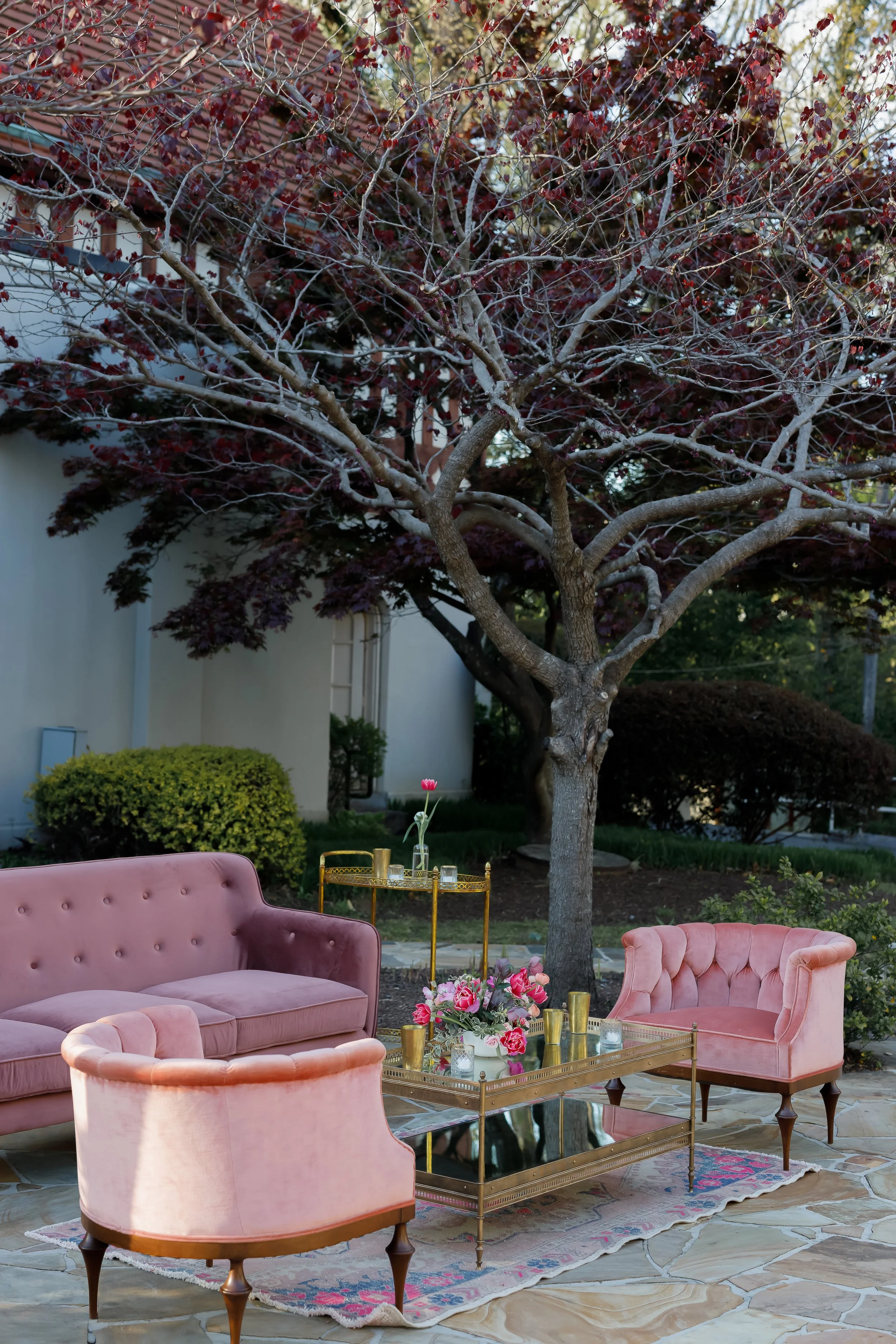 A cozy outdoor patio area with pink velvet furniture including a sofa and two armchairs, a glass-topped coffee table with floral decorations, in front of a large leafless tree and a house with greenery in the background.
