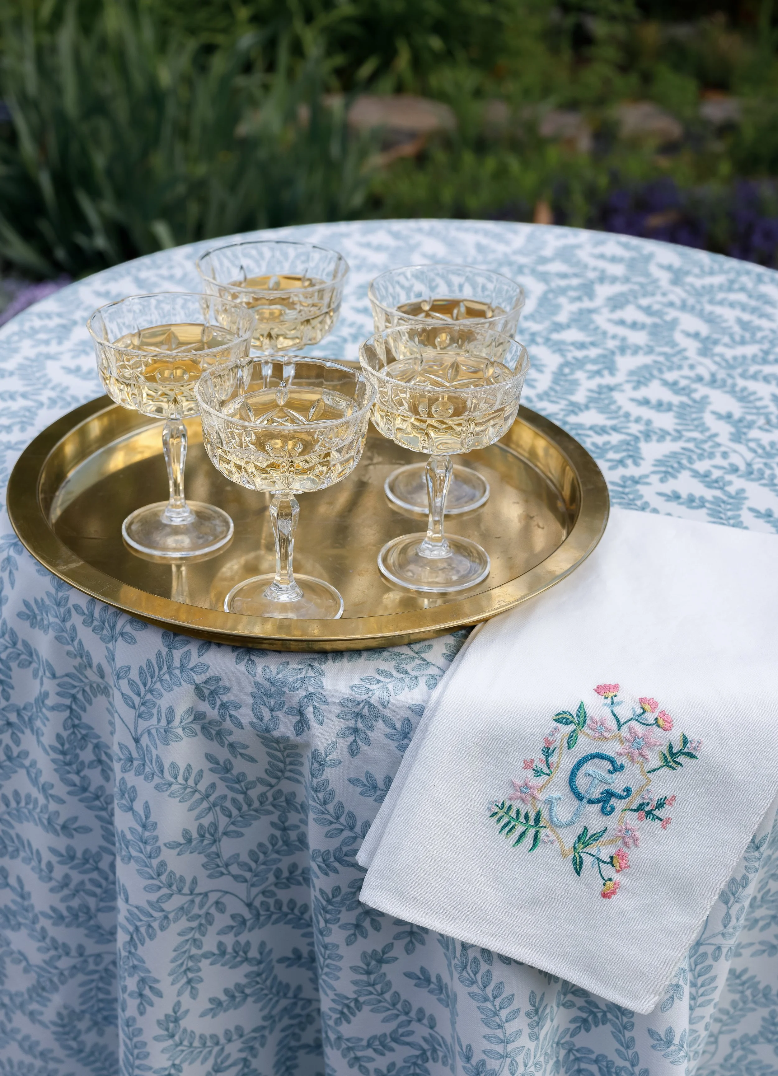 Six crystal cocktail glasses filled with a light-colored drink on a round brass tray, placed on a white tablecloth with blue leaf patterns, outdoors with greenery in the background, and a white embroidered napkin with a monogrammed letter 'G' and flo