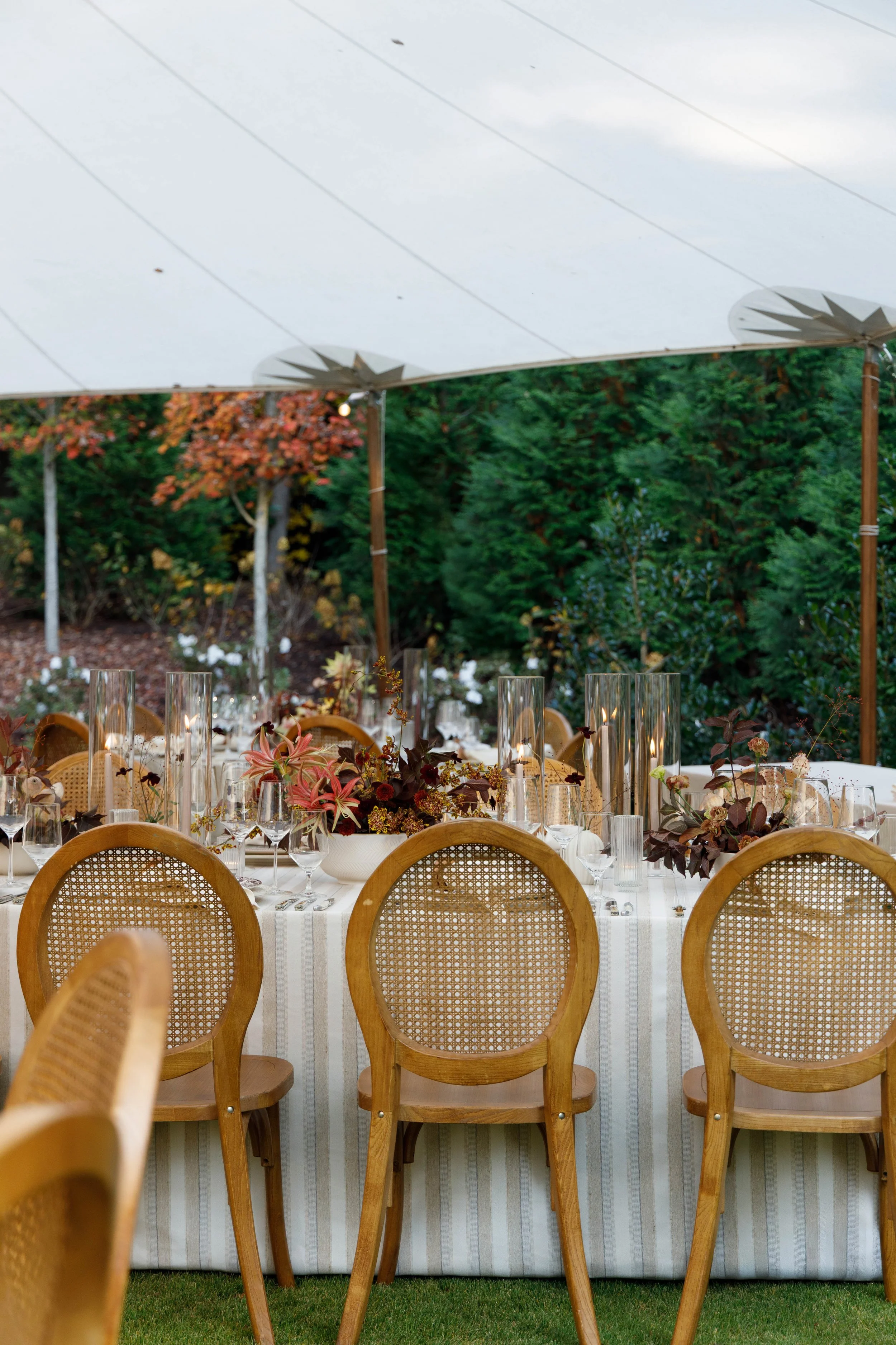 Elegant outdoor dining setup under a white canopy with a long table decorated with floral centerpieces, tall glass candle holders, and wooden chairs, surrounded by lush greenery and trees.
