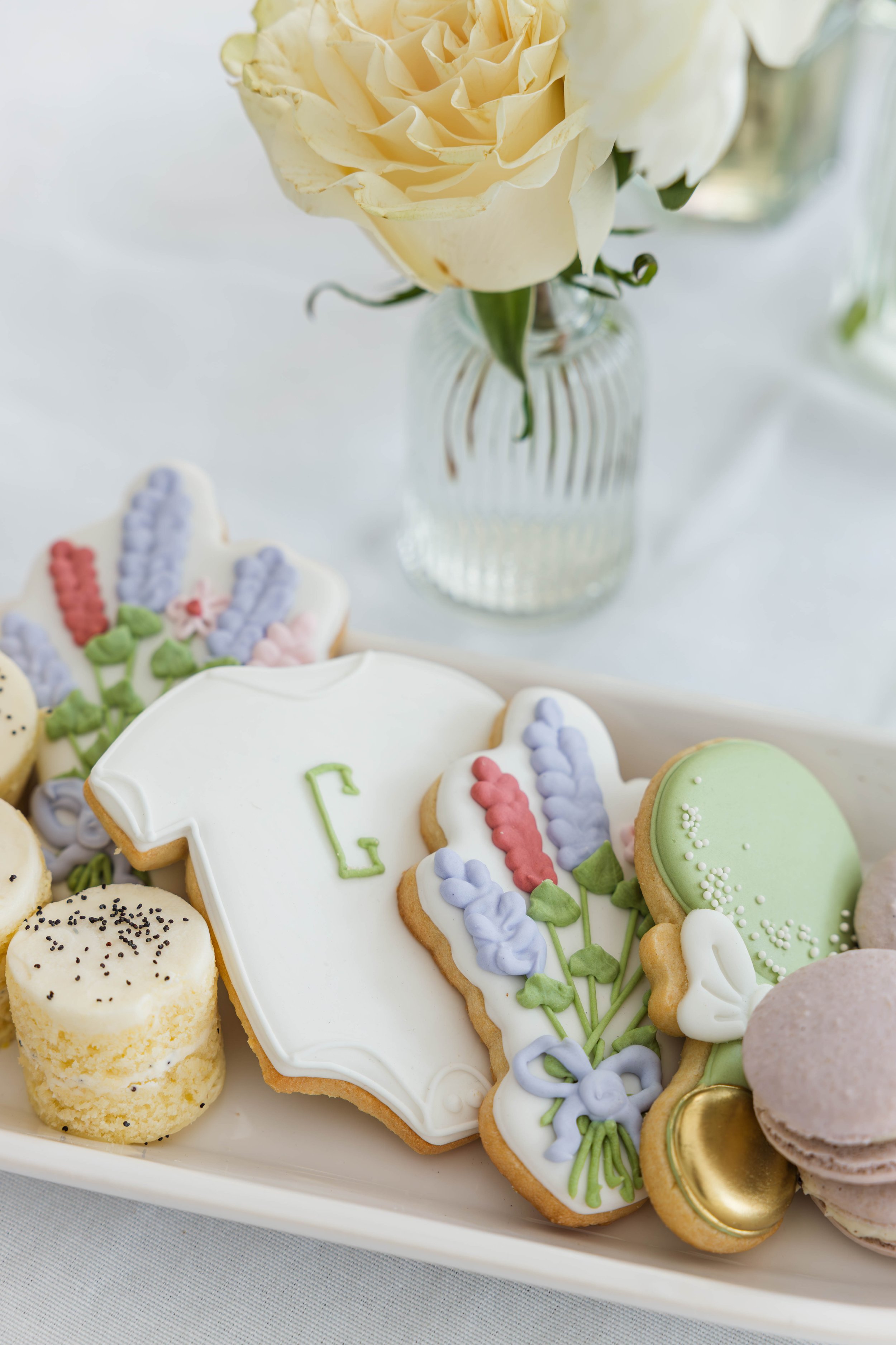 Assorted decorated cookies on a white tray, with a vase of cream-colored roses in the background.