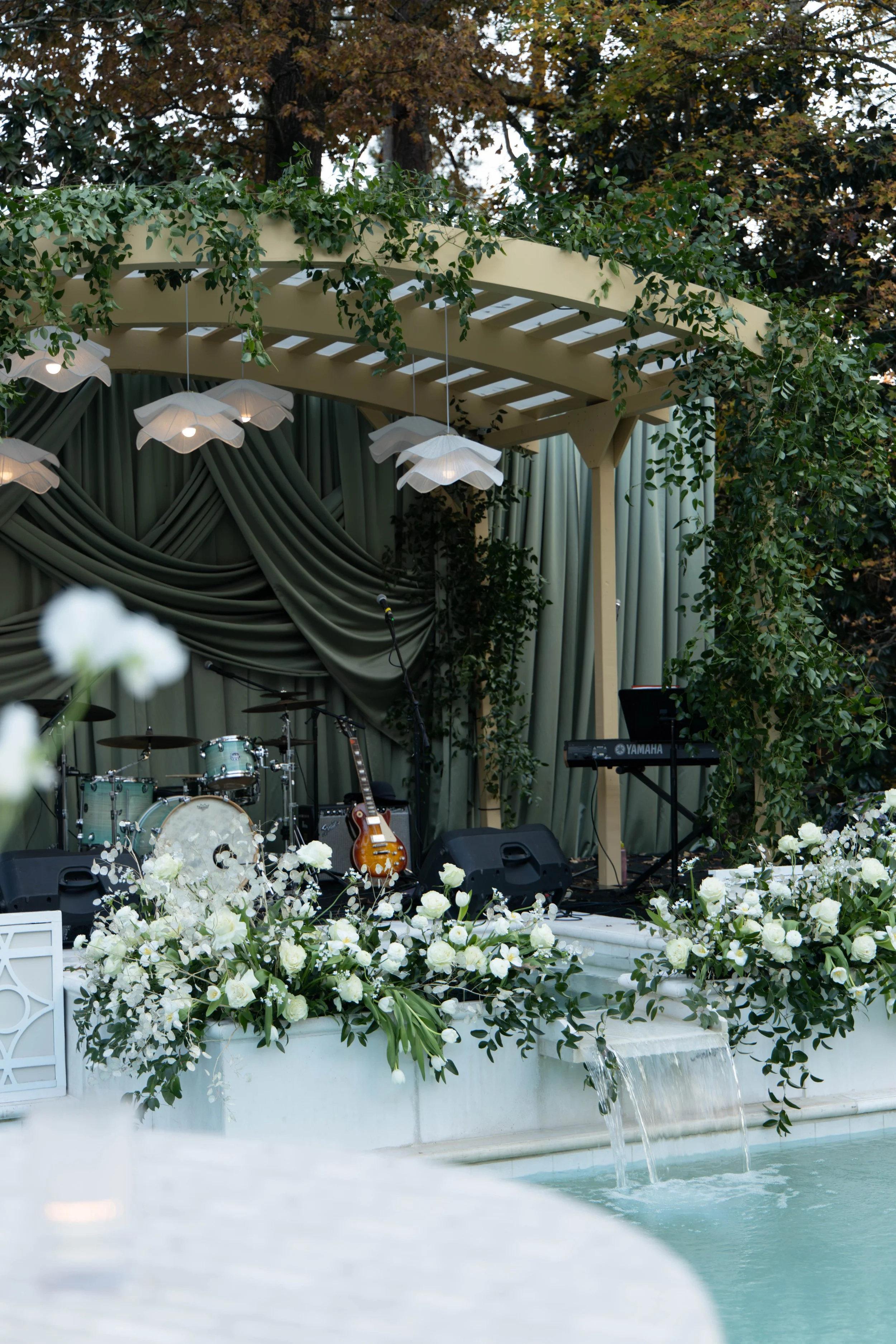 Stage setup with musical instruments, microphones and speakers, decorated with white flowers and greenery, near a pool with water cascading in front.