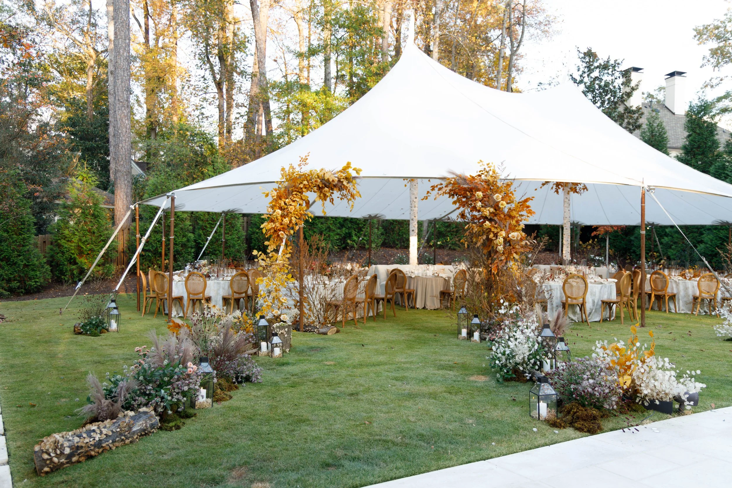 Outdoor party setup with a large white tent, decorated with autumn-themed floral arrangements and candles on the grass, surrounded by trees.