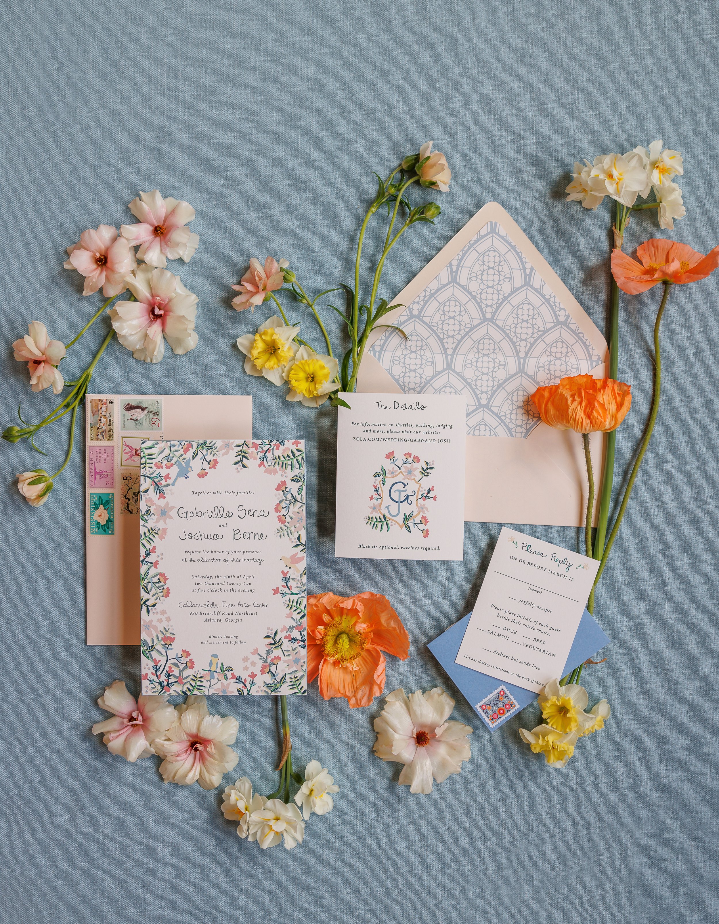Wedding invitation suite with floral accents on a blue fabric background, including invitations, RSVP card, details card, and colorful flowers.