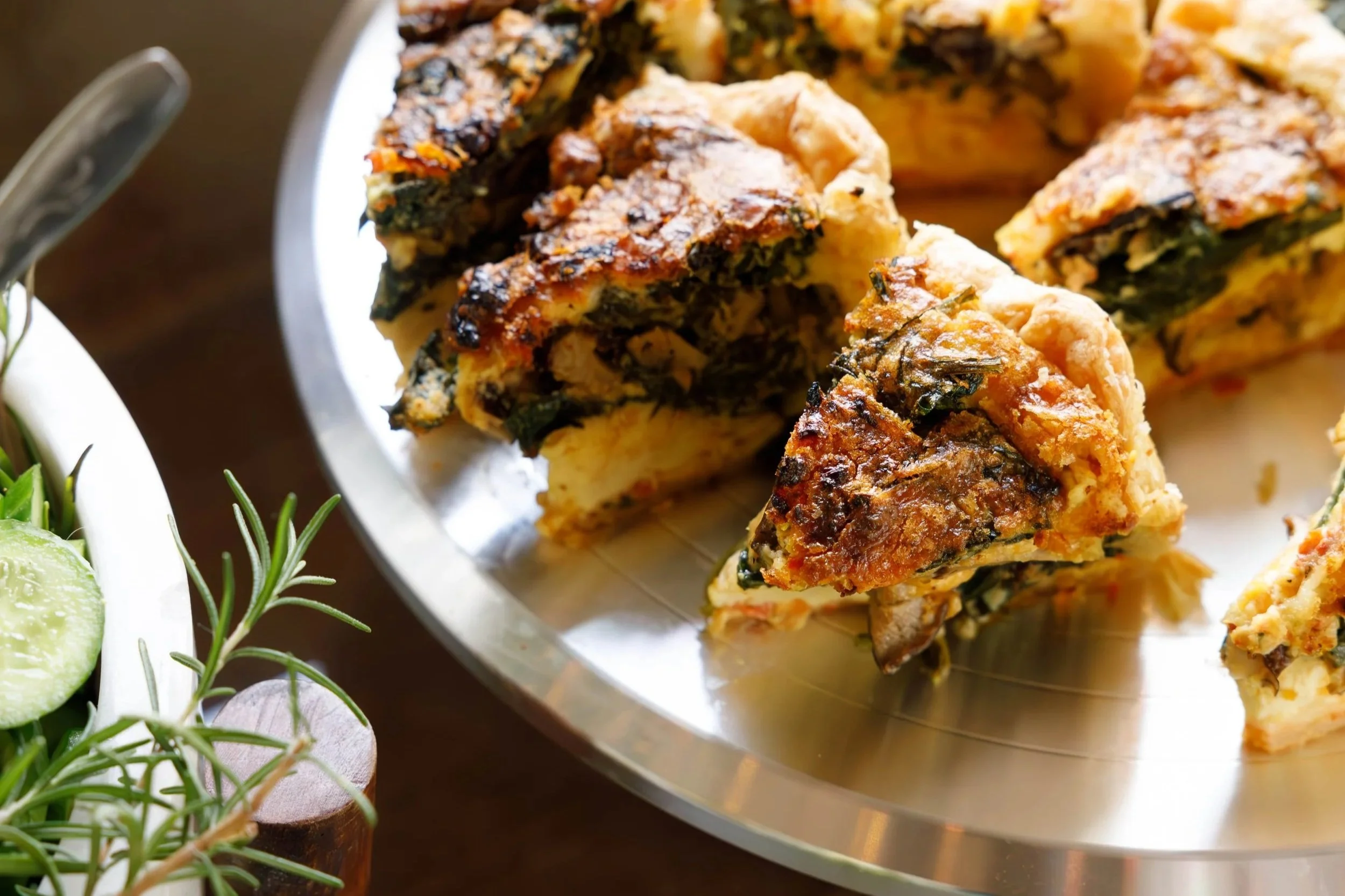 Slices of spinach and cheese frittata on a silver serving platter, with a side of cucumber and rosemary garnishing in a white bowl.